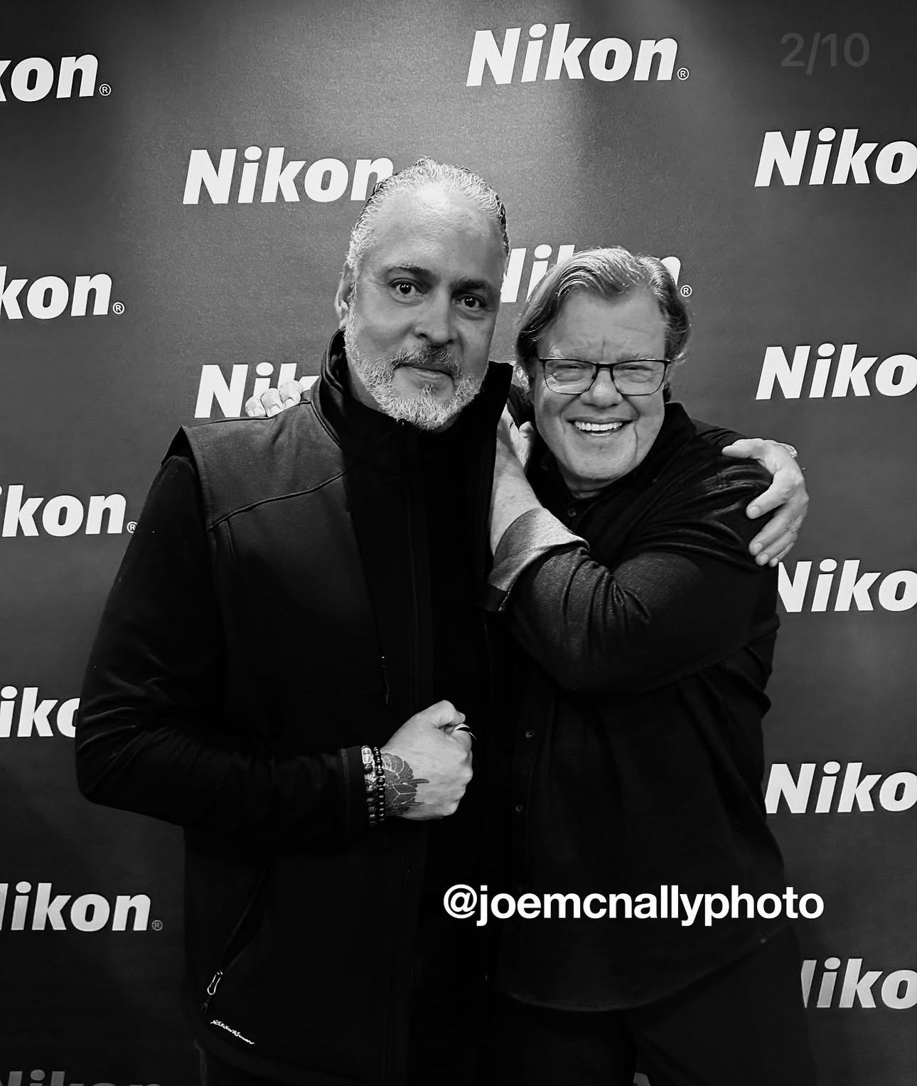 Two people pose together against a Nikon branded backdrop in black and white.