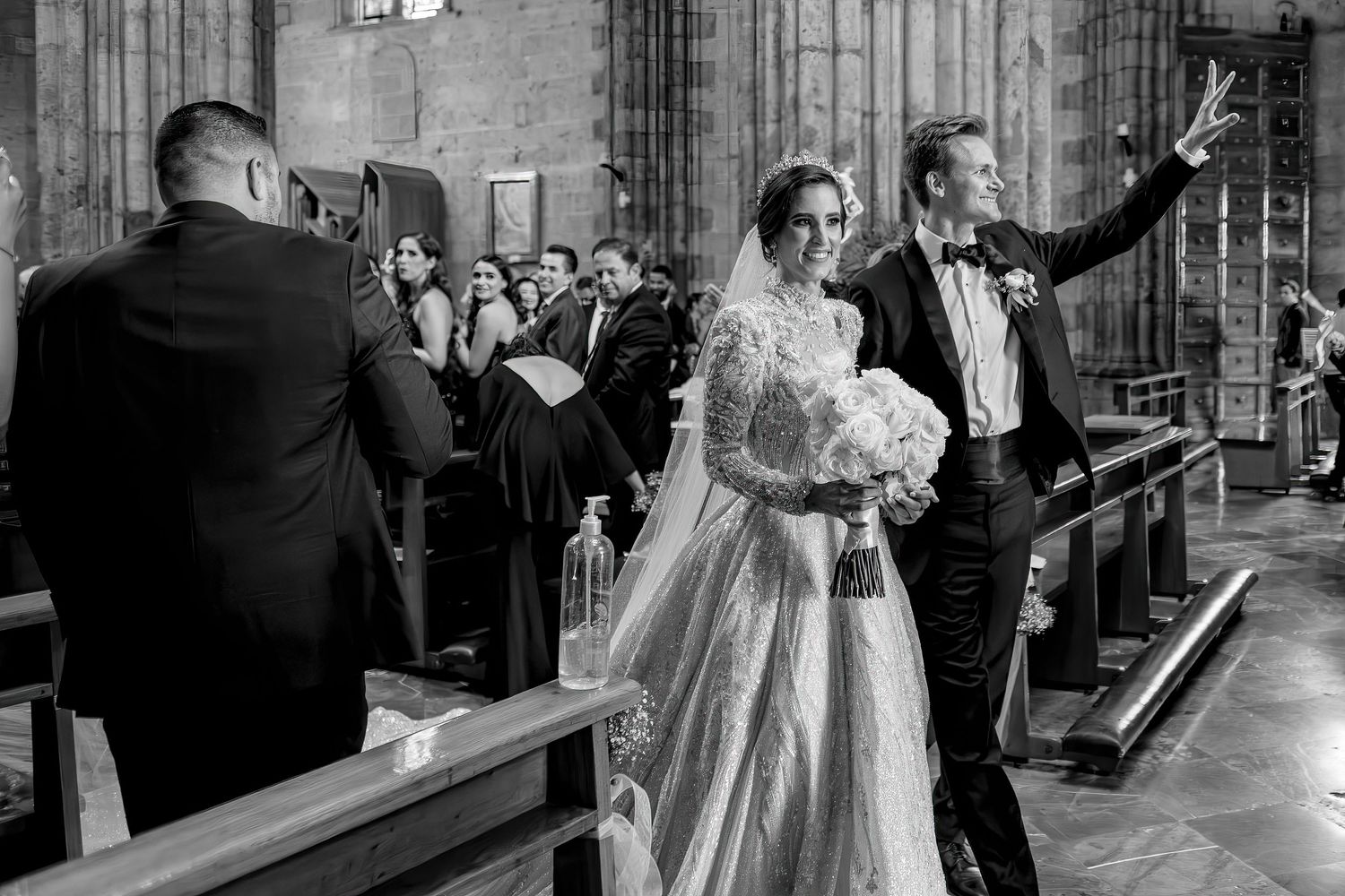 Black and white wedding photo of bride and groom walking down church aisle, groom waving to guests.
