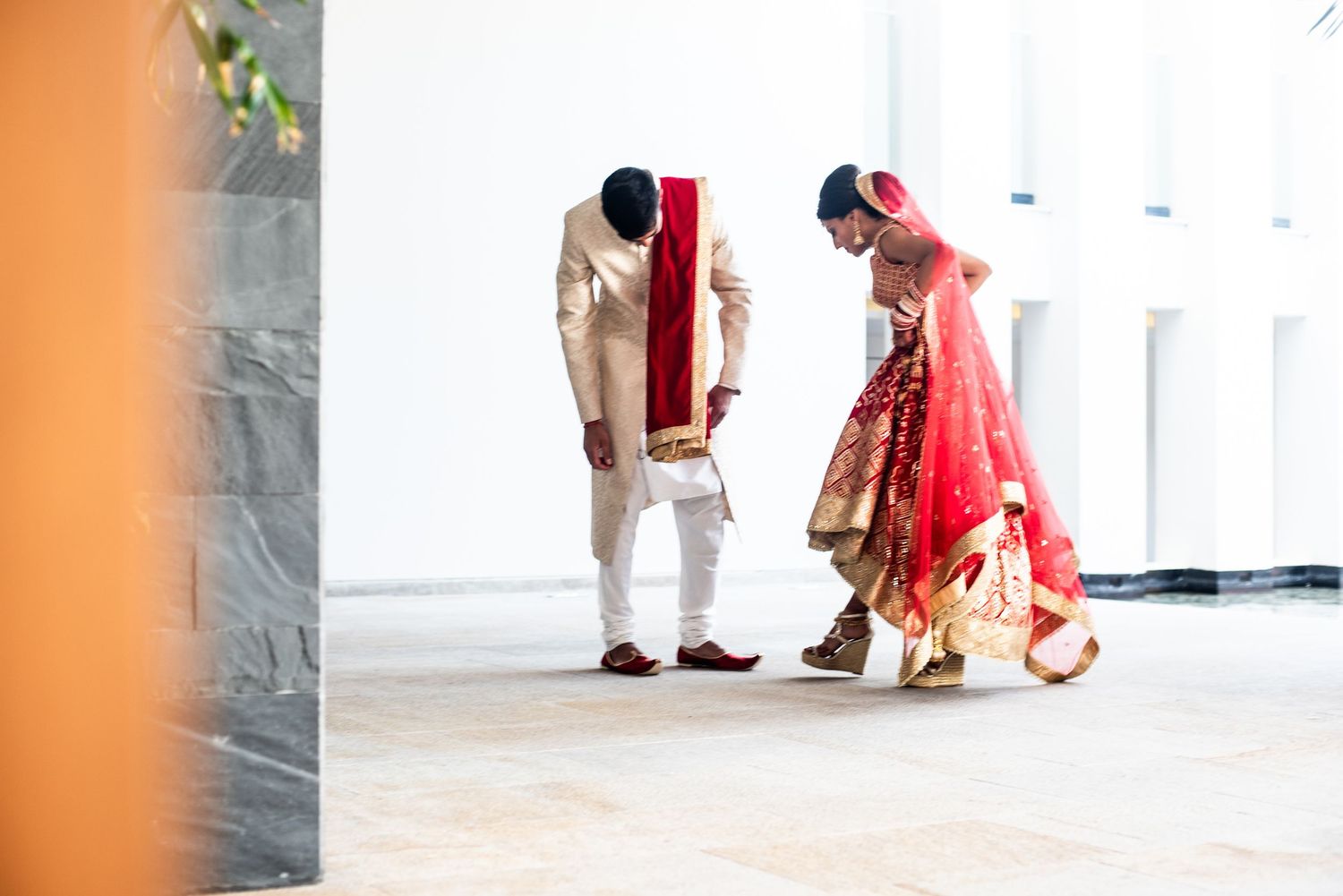Indian bride in red lehenga and groom in cream sherwani walk together during traditional wedding ceremony.