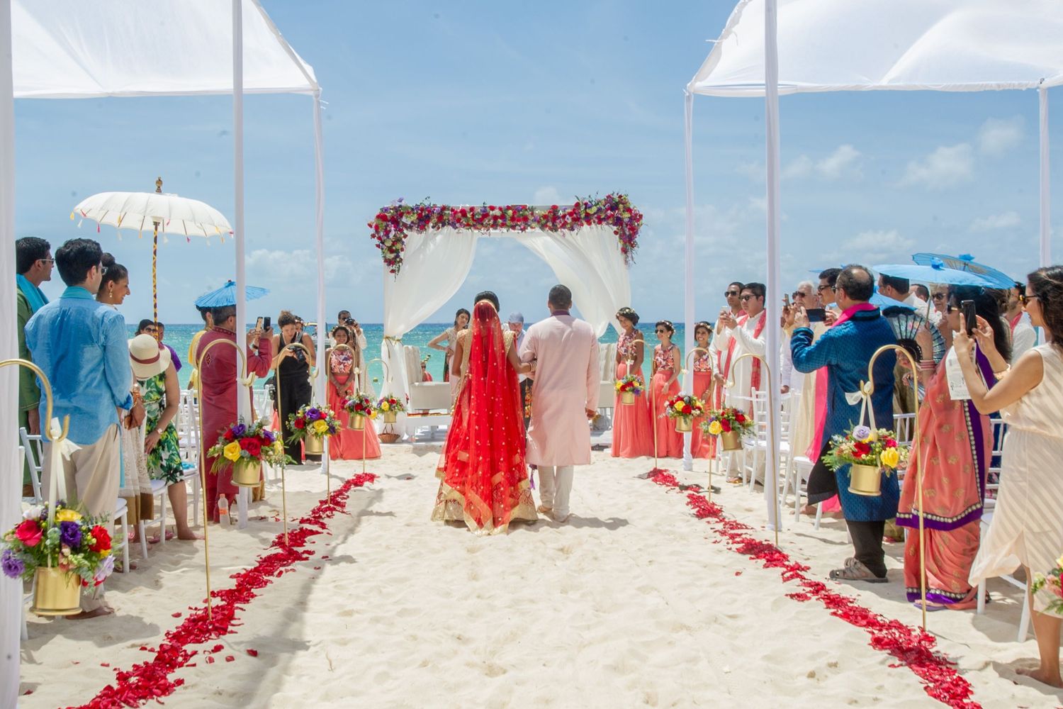 Couple walks down rose petal aisle at elegant beach wedding ceremony with floral arch and guests in colorful attire.