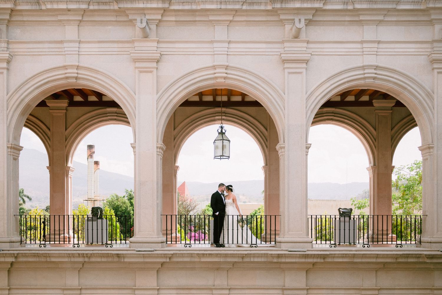 Bride and groom share a romantic moment under elegant arched colonnade with scenic mountain views at luxury wedding venue.