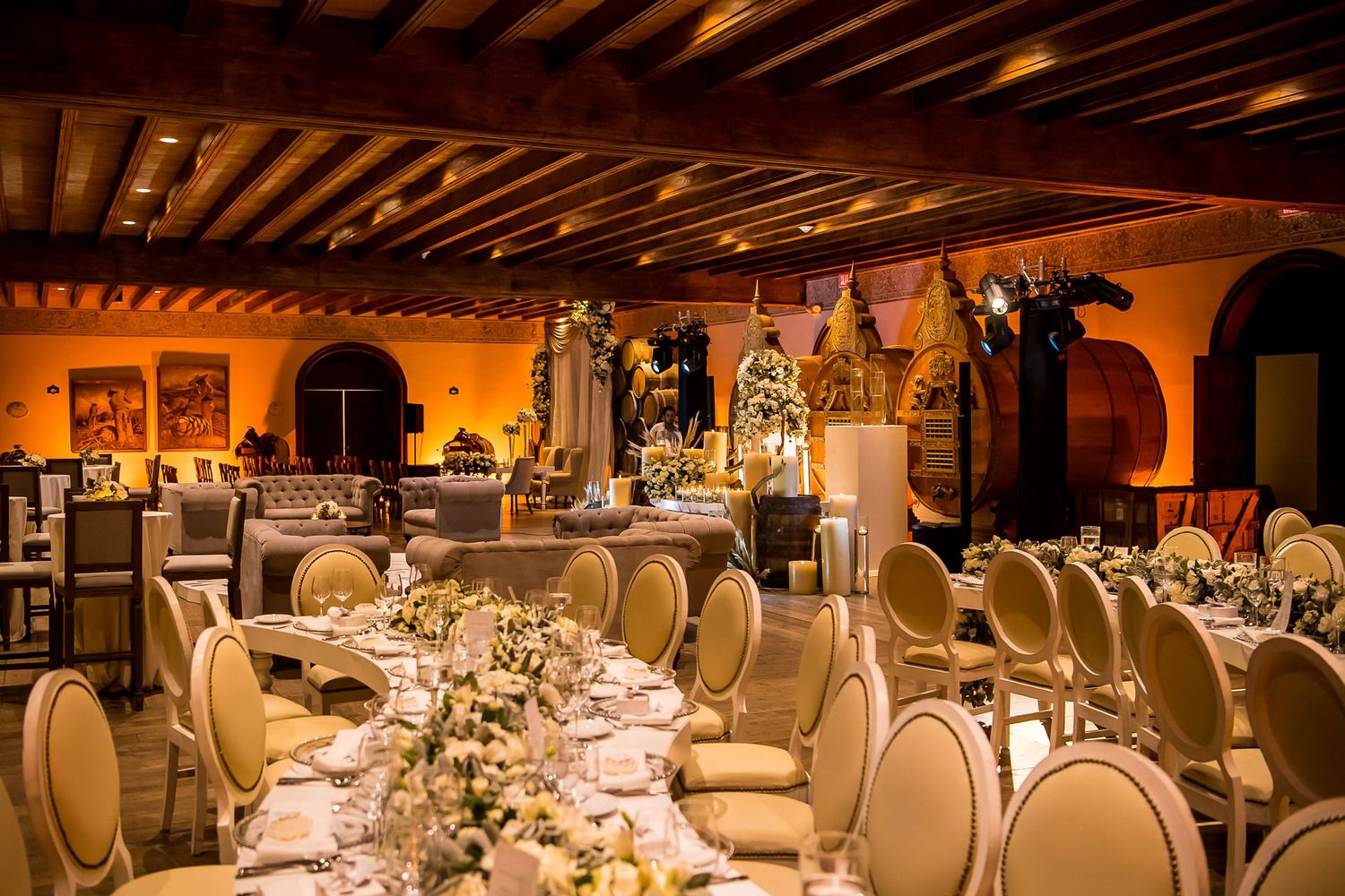 Elegant wedding reception hall with ivory chairs, floral centerpieces, and warm golden lighting in a rustic venue.