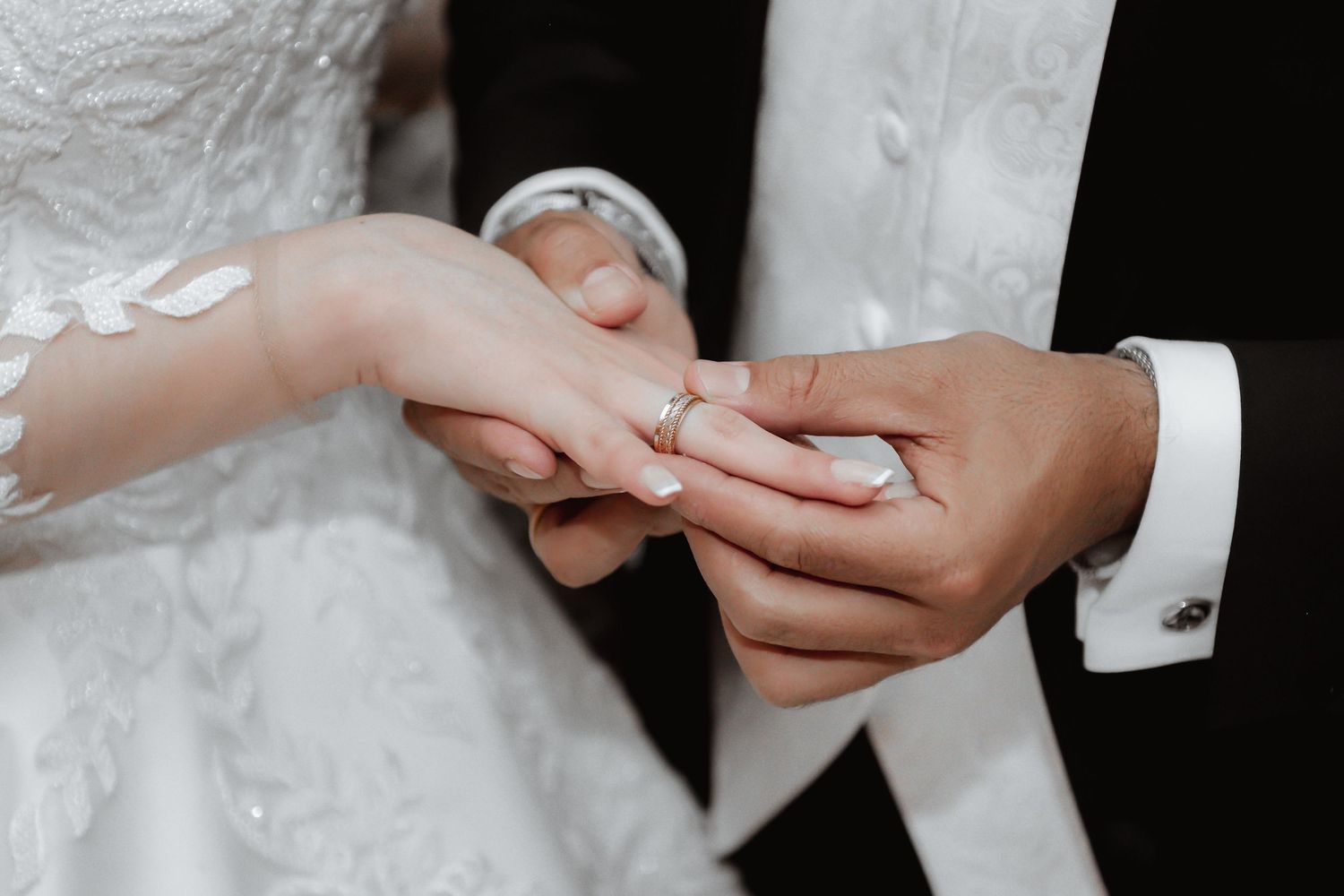 Close-up of hands during wedding ring exchange ceremony showing delicate lace sleeve detail.
