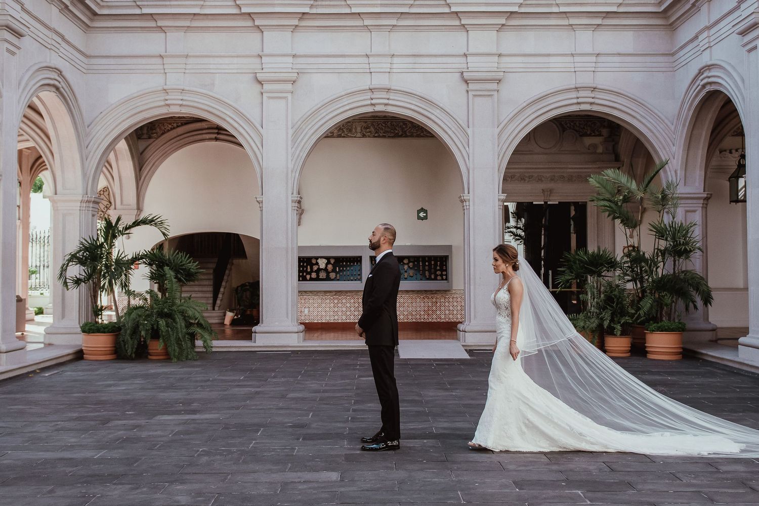 Bride and groom share a first look moment in an elegant arched courtyard with white columns and lush tropical plants.