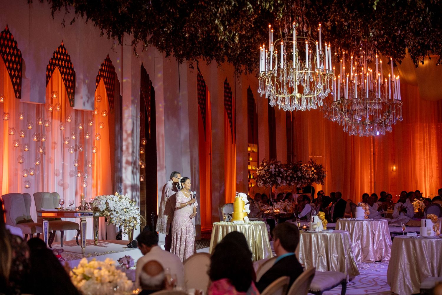 Elegant wedding reception with chandelier, orange drapes, couple sharing first dance, floral arrangements, and candlelit tables.