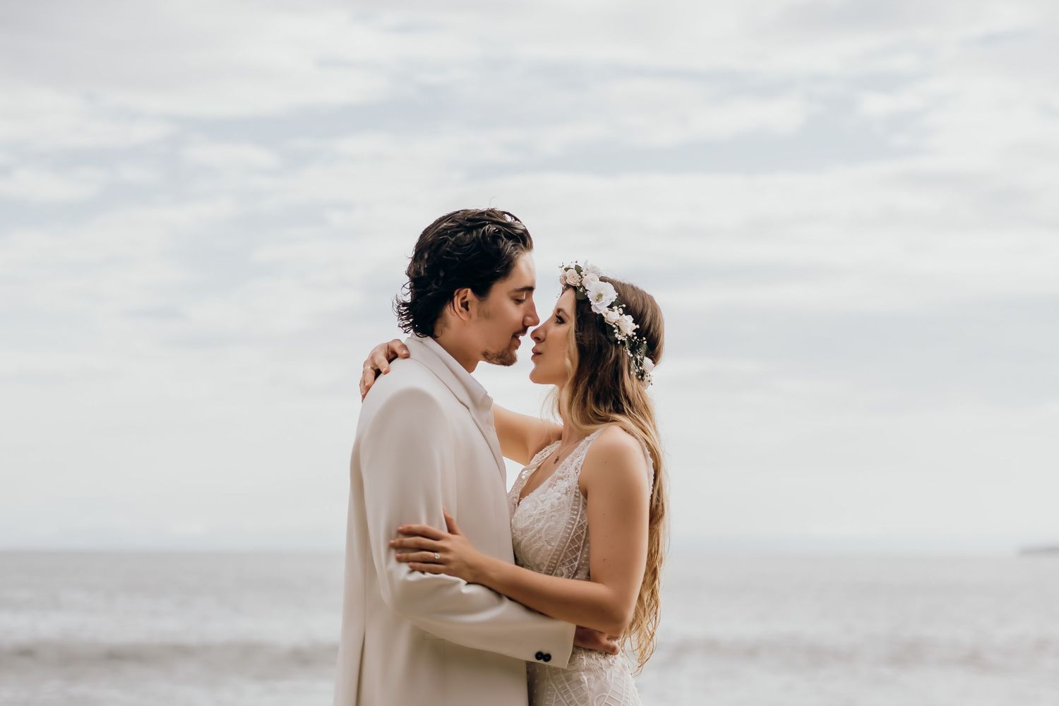 A bride and groom share a romantic moment on a beach, with the bride wearing a floral crown and the groom in a white suit.