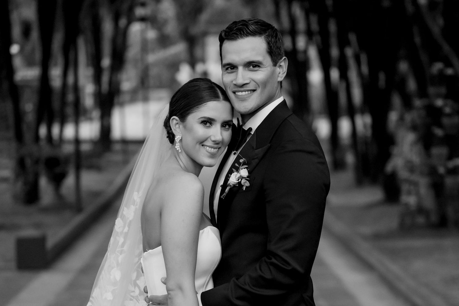 Black and white wedding portrait of a smiling bride and groom posing together outdoors along a tree-lined path.