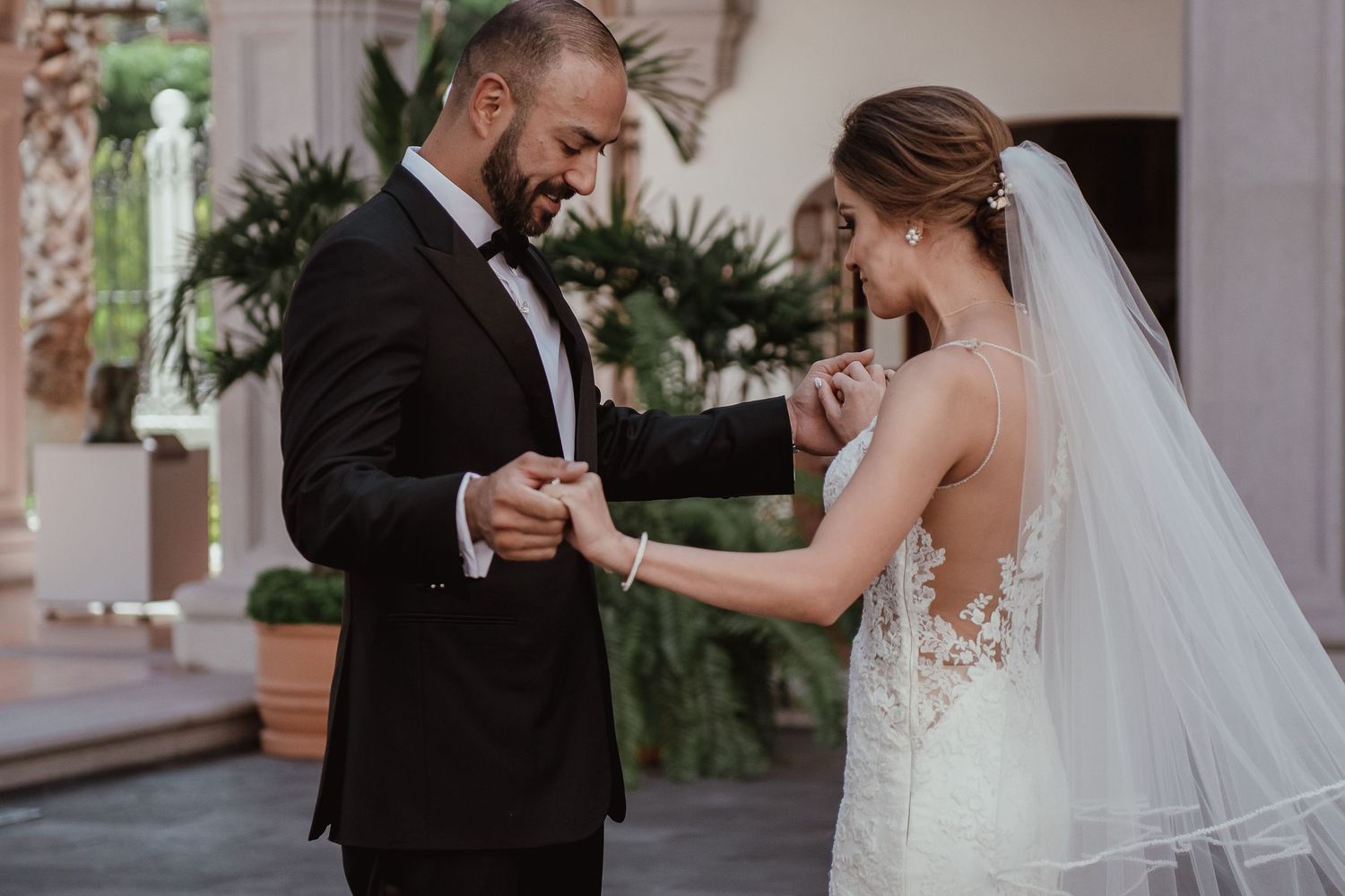 A groom in a black tuxedo holds his bride's hand as she wears a lace backless wedding gown and veil.