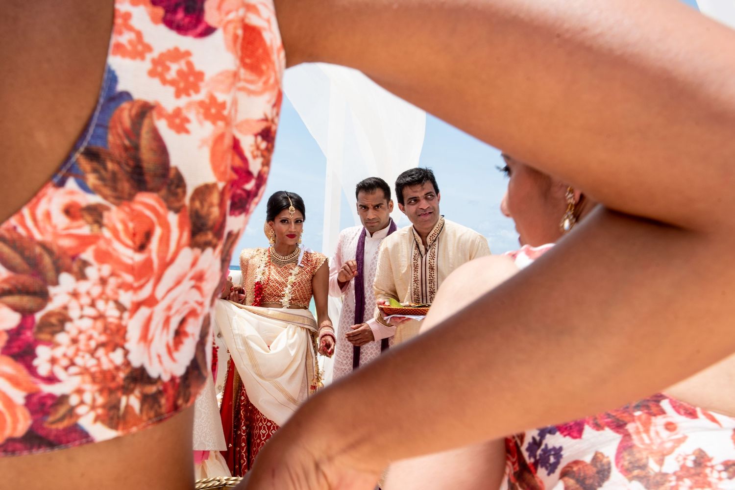 Indian wedding couple in traditional attire walking down the aisle, framed by guests in floral dress.