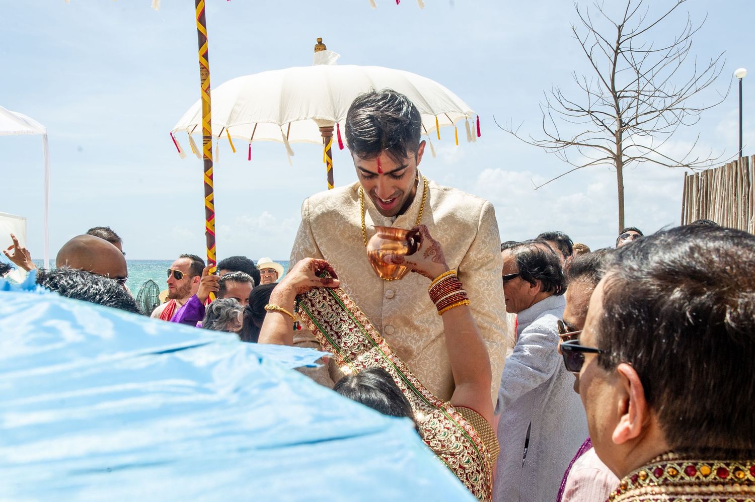 Groom in traditional Indian sherwani bowing during outdoor wedding ceremony with guests and white umbrella.