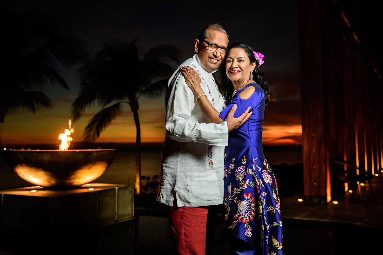 Couple embracing at tropical sunset venue with palm trees and fire feature in background.