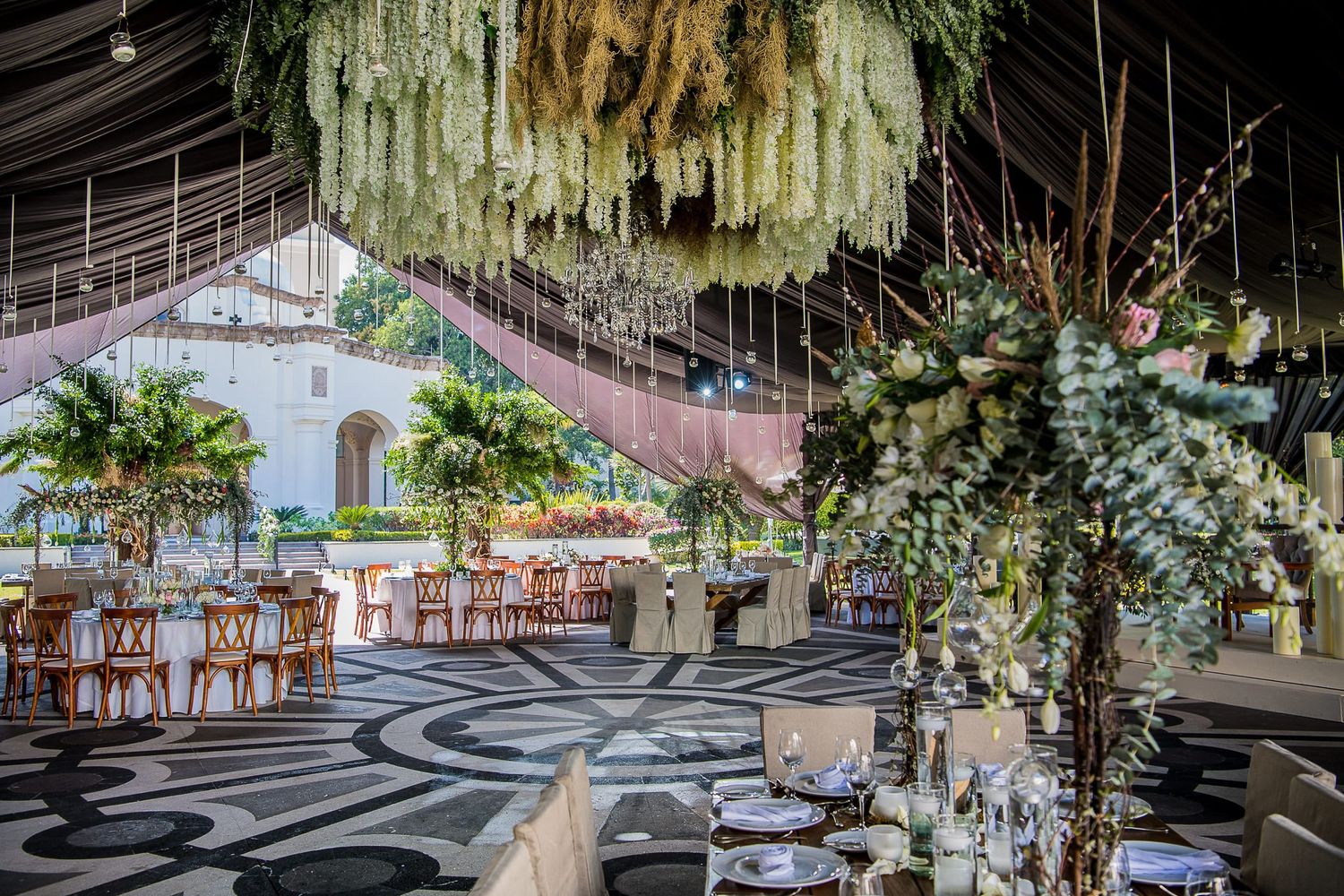 Elegant outdoor wedding reception venue with floral ceiling installations, palm trees, and decorated tables under a canopy.