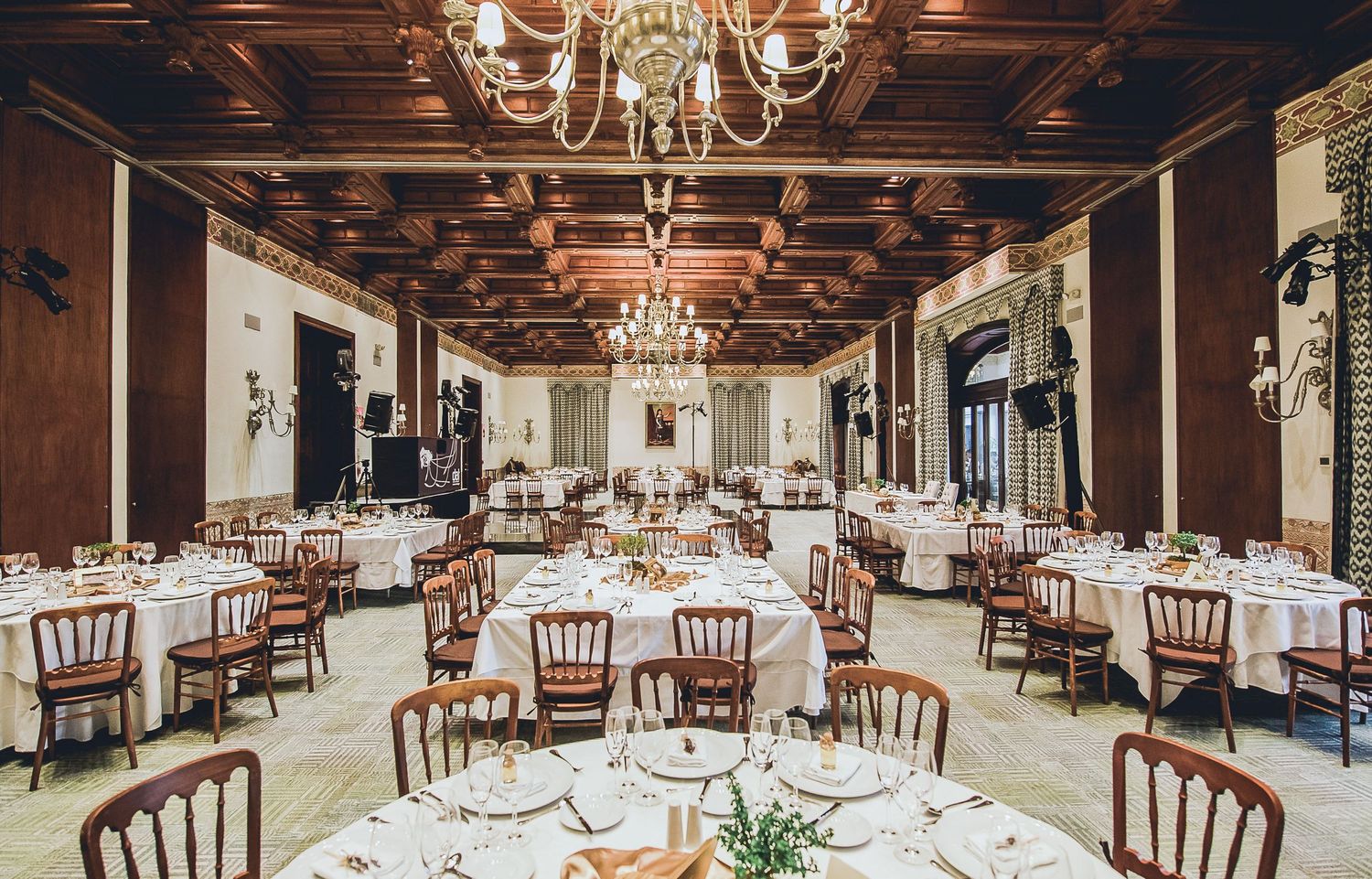 Elegant banquet hall with chandeliers, wood-paneled ceiling, and round tables set for a formal dining event.