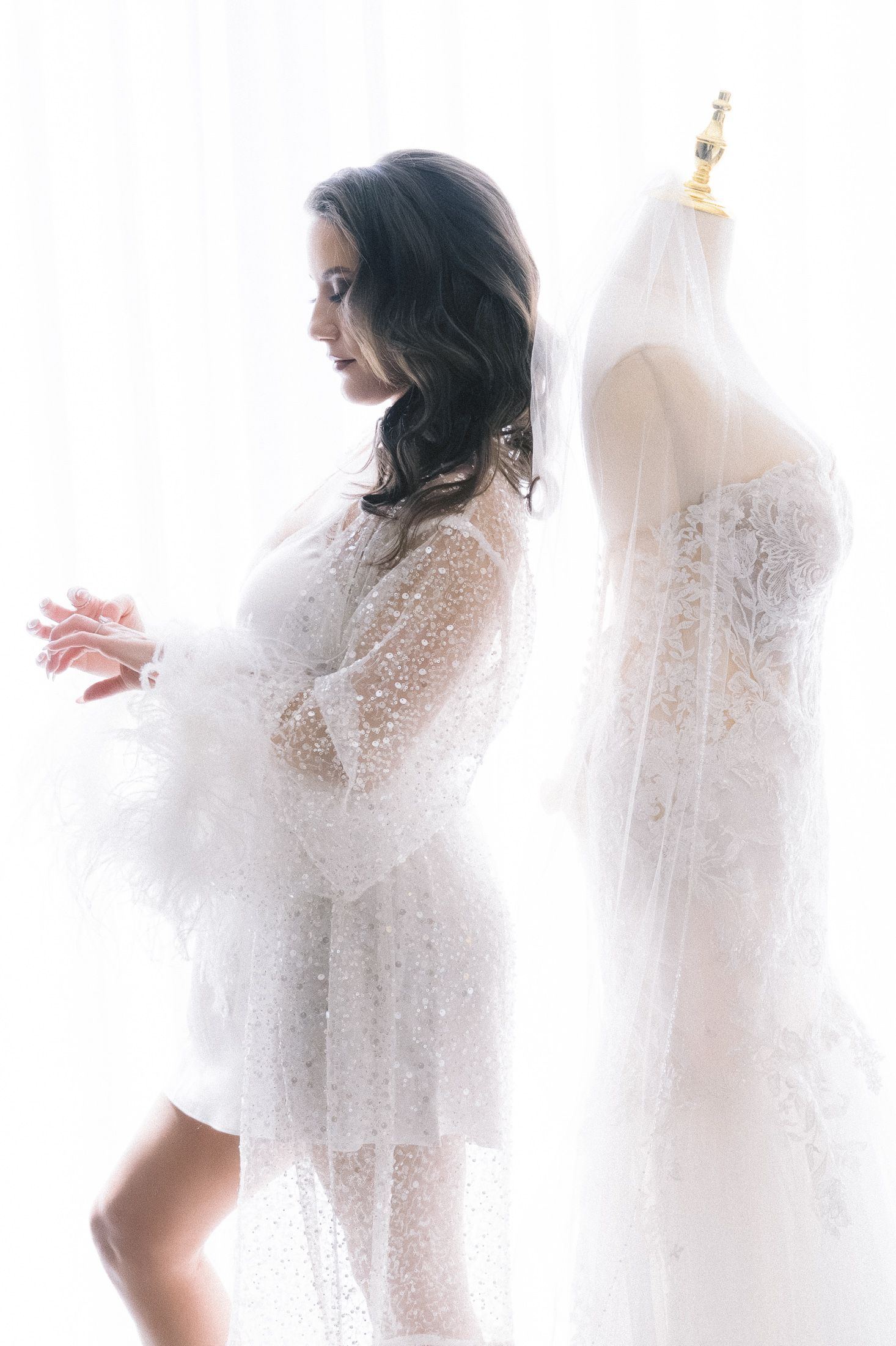 Ethereal silhouette of white lace bridal robe and wedding dress on mannequin in soft window light.
