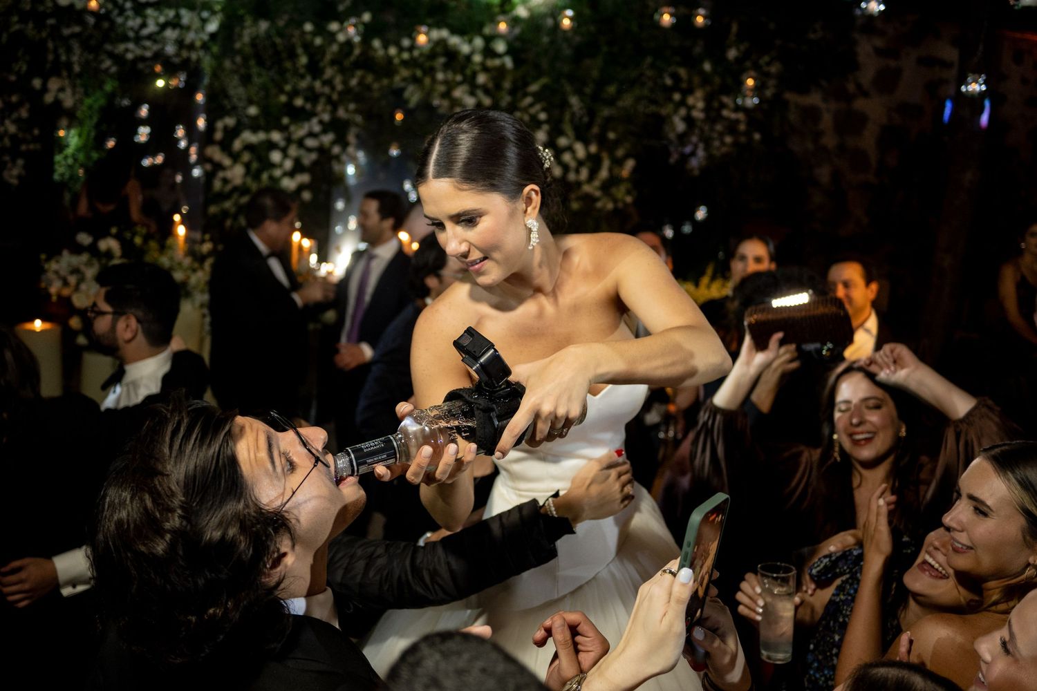 Bride in strapless gown pours champagne for guests at elegant outdoor wedding reception at night.