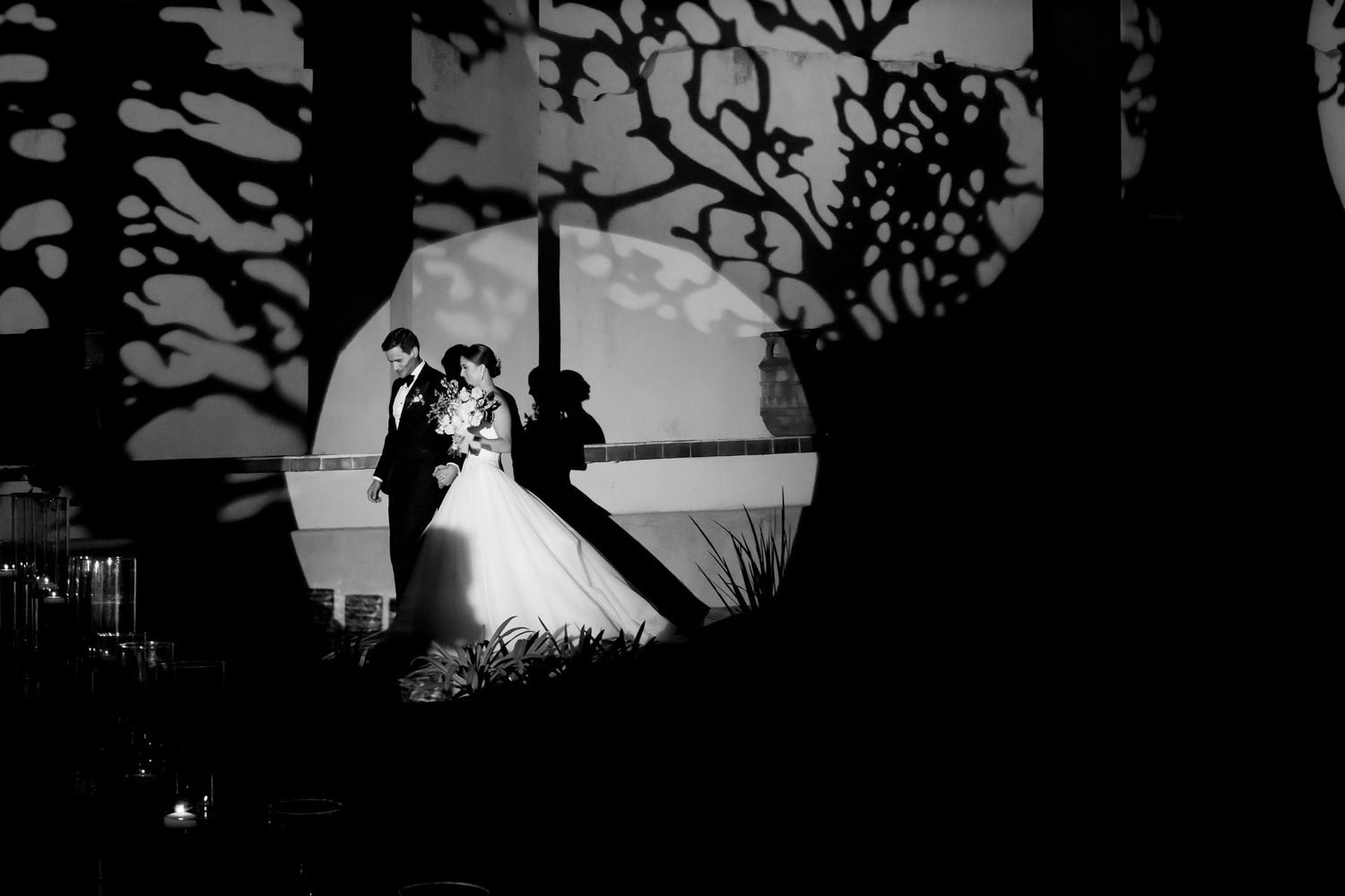 Black and white silhouette of bride and groom embracing under decorative tree shadows at night.