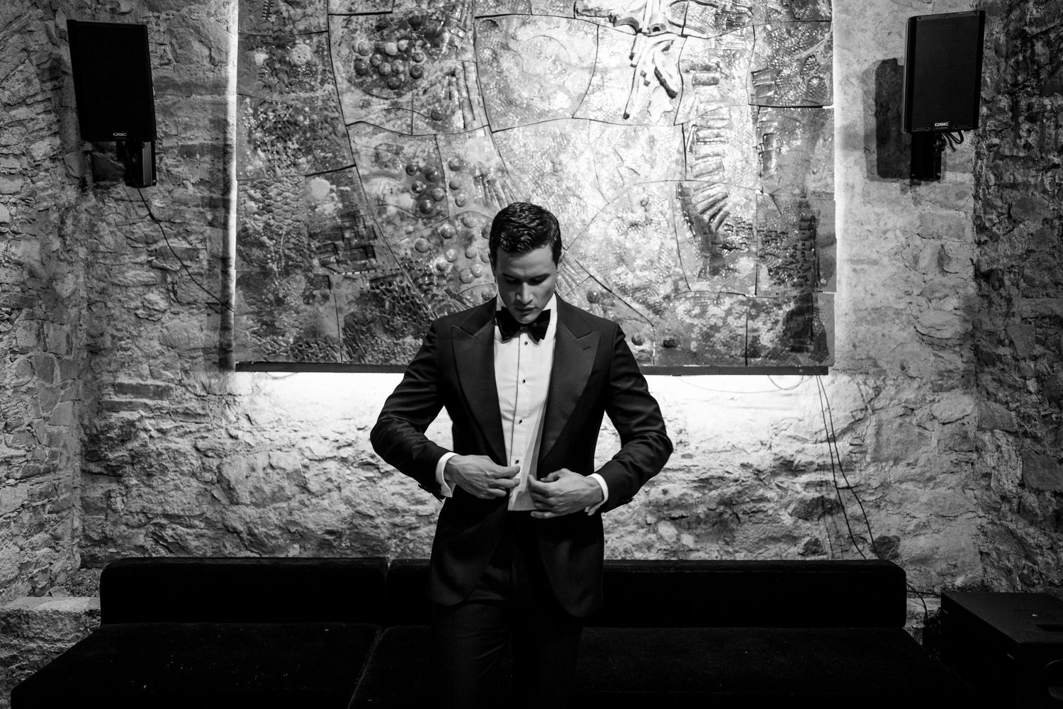 Black and white photo of a man in a tuxedo adjusting his jacket in an elegant stone-walled venue.