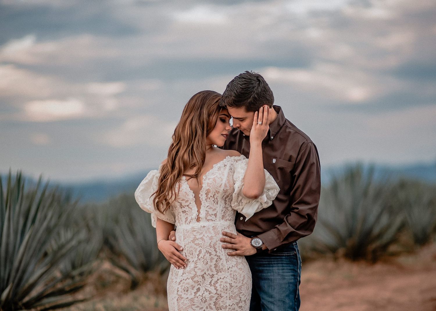 Couple in romantic embrace among agave plants, woman in lace dress, man in brown shirt, cloudy sky backdrop.