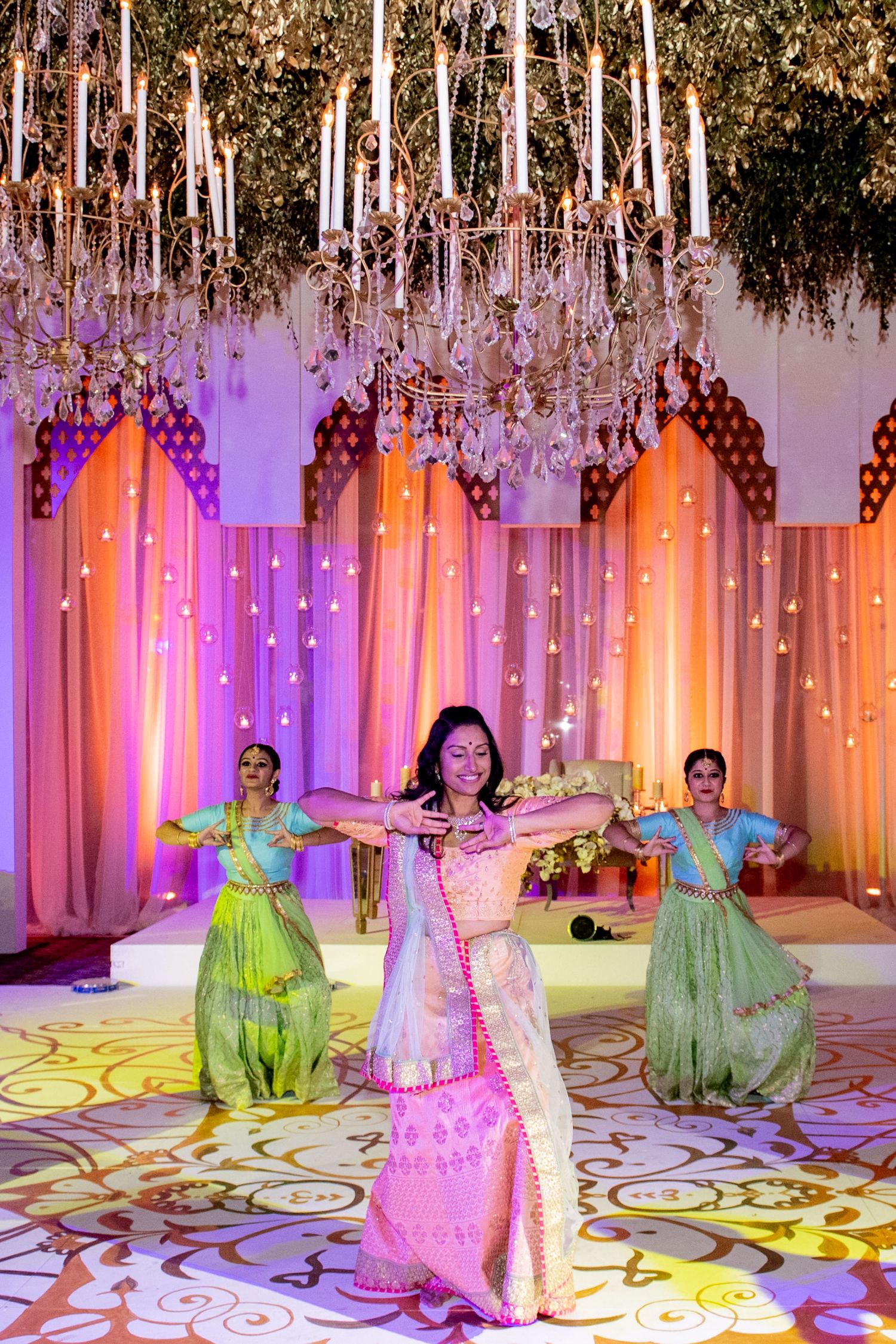 Three women in colorful sarees perform a traditional Indian dance under a crystal chandelier at a vibrant wedding celebration.