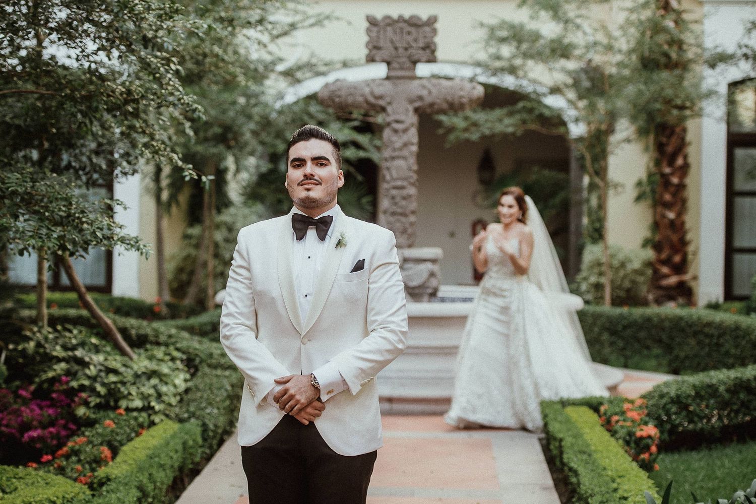 Groom in white tuxedo stands in garden path as bride in white gown approaches during wedding first look.
