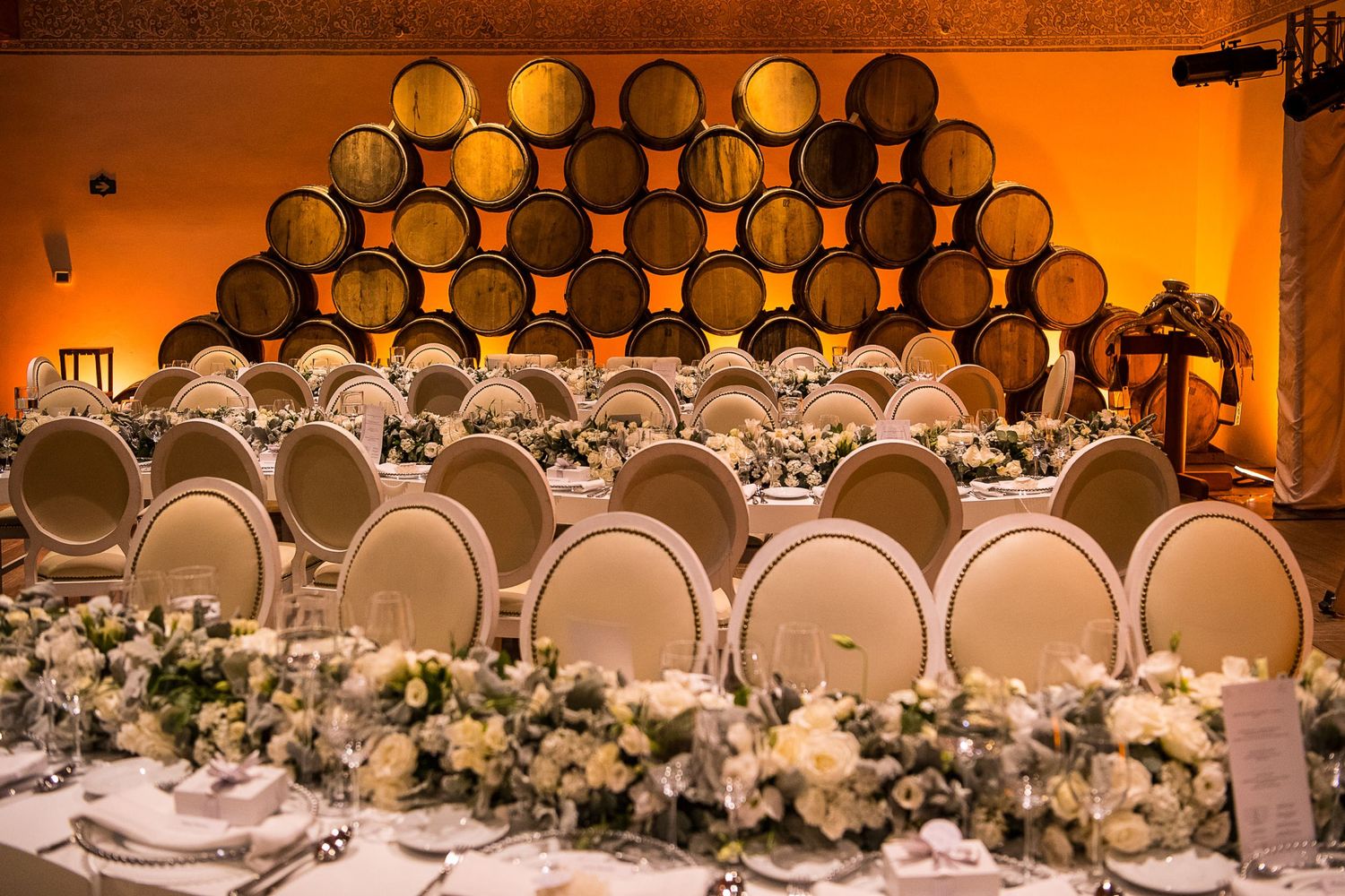 Elegant winery wedding reception with barrel wall backdrop, ivory oval chairs, and white floral centerpieces.