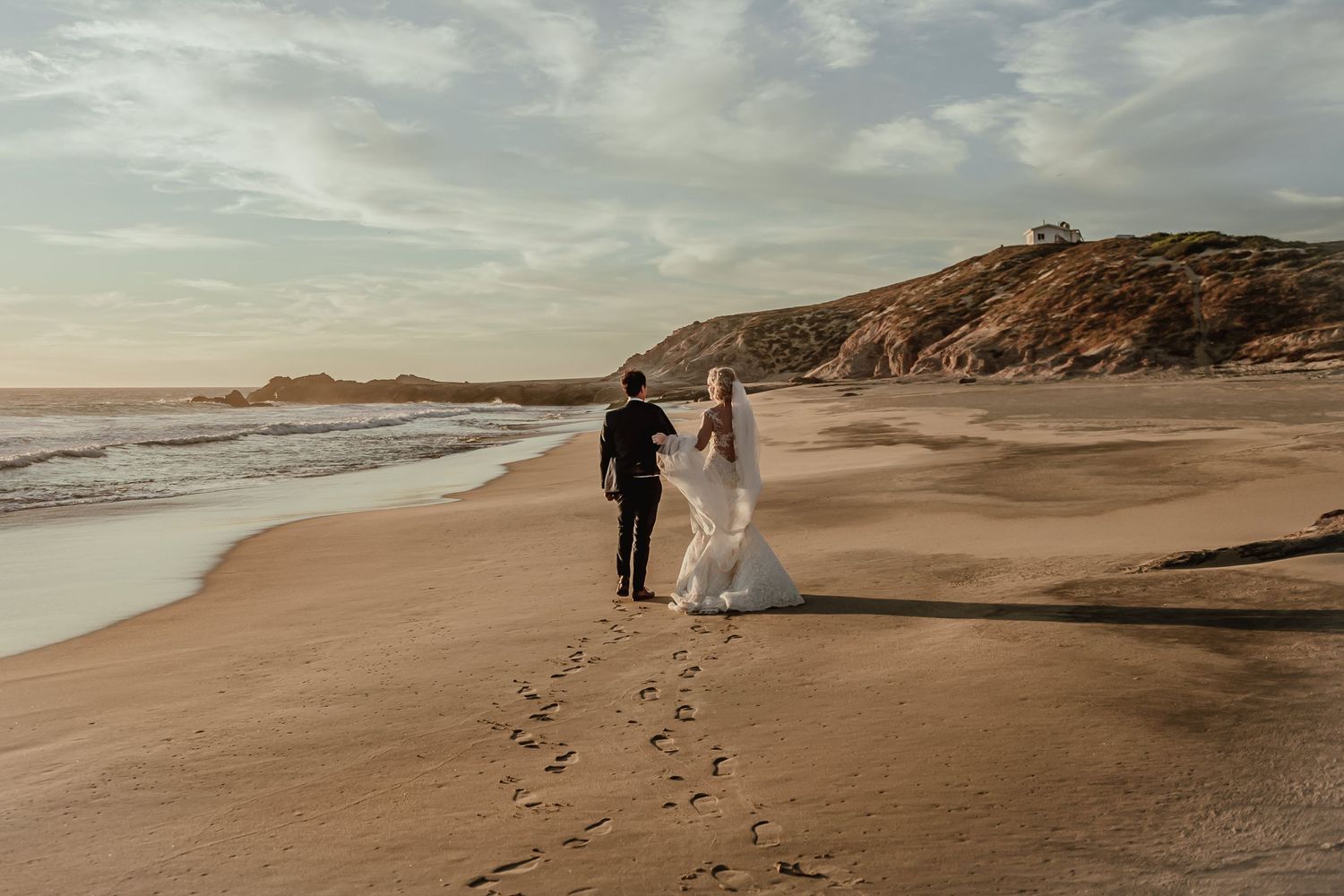 Newlyweds walk hand in hand on a scenic beach at sunset leaving footprints in the sand.