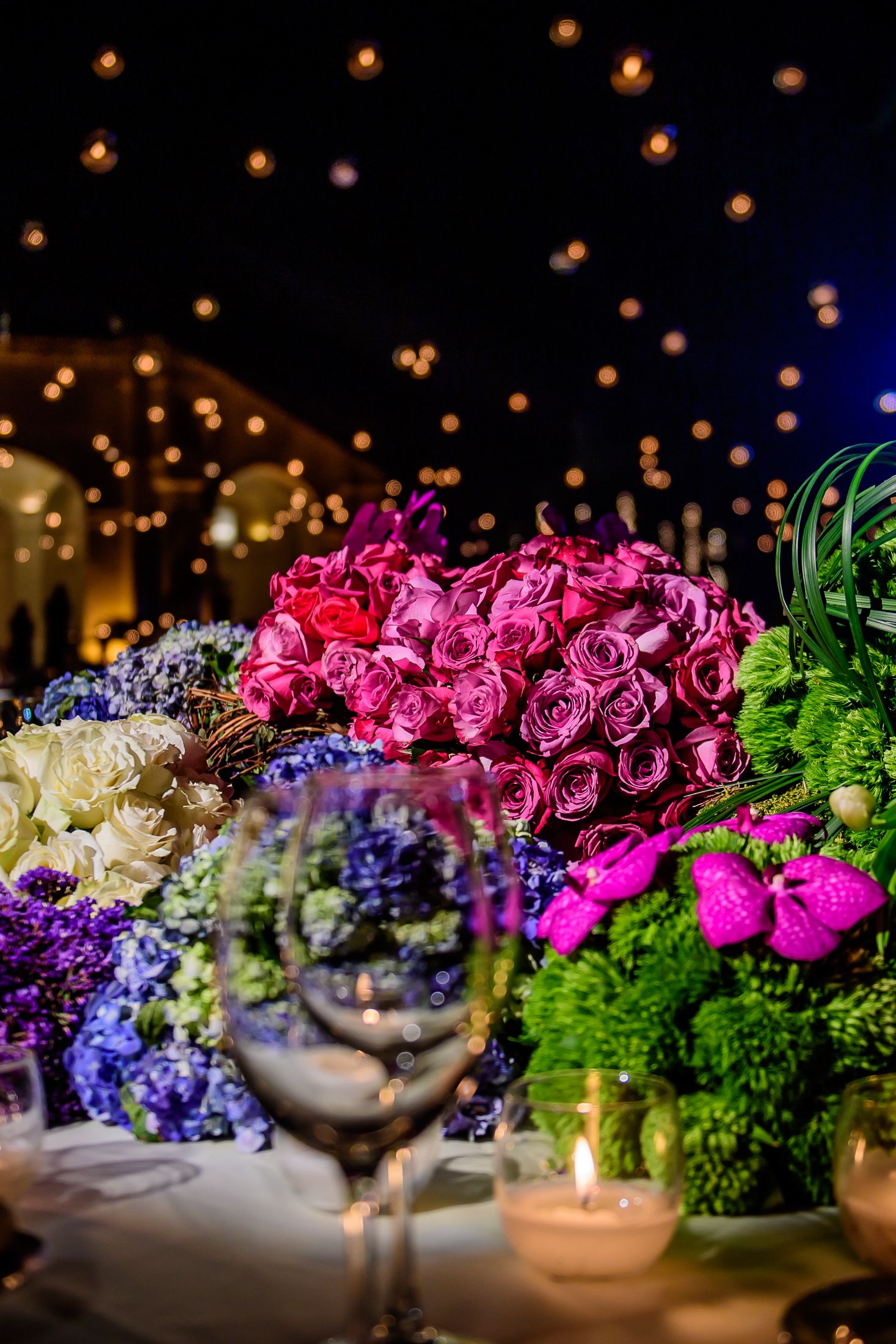 Colorful floral centerpiece featuring pink roses, purple blooms, and greenery with glowing lanterns above.