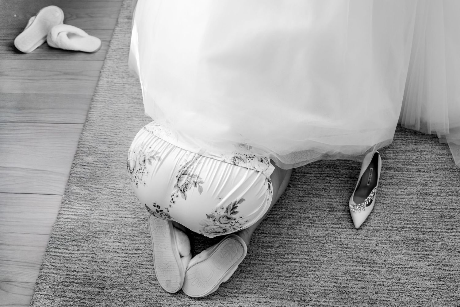 Black and white photo of bride kneeling on floor being helped into wedding shoes by bridesmaid.