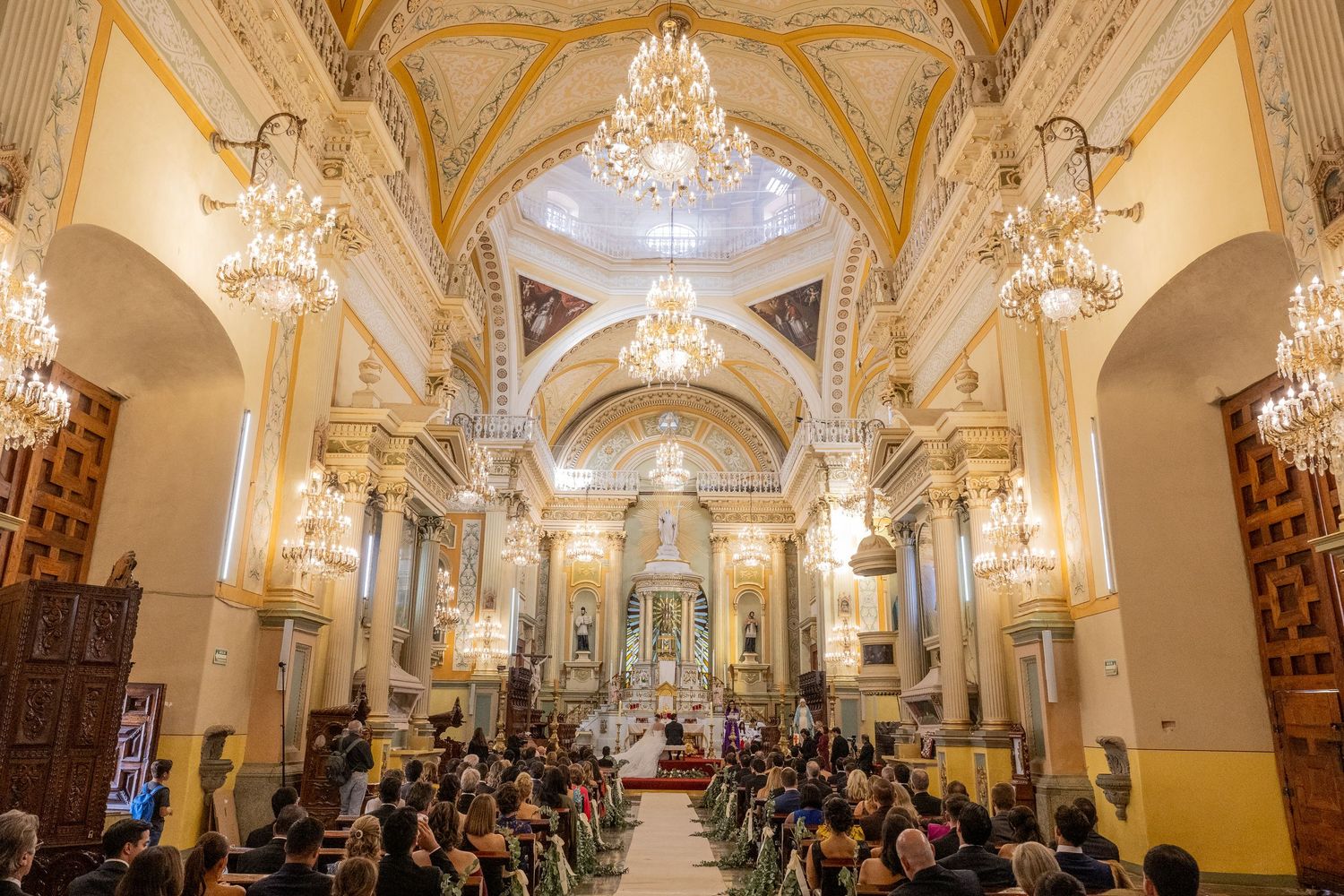 Elegant church wedding ceremony with ornate golden decor, chandeliers, and guests seated in pews beneath a grand vaulted ceiling.