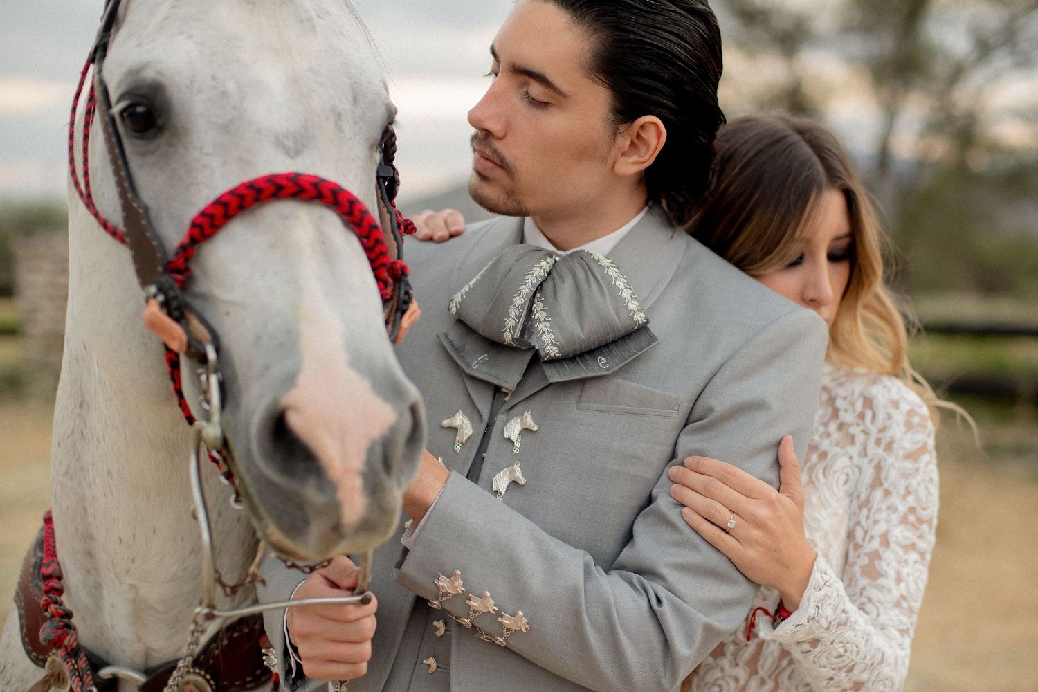 Couple in traditional charro attire posing with a white horse with red bridle outdoors.