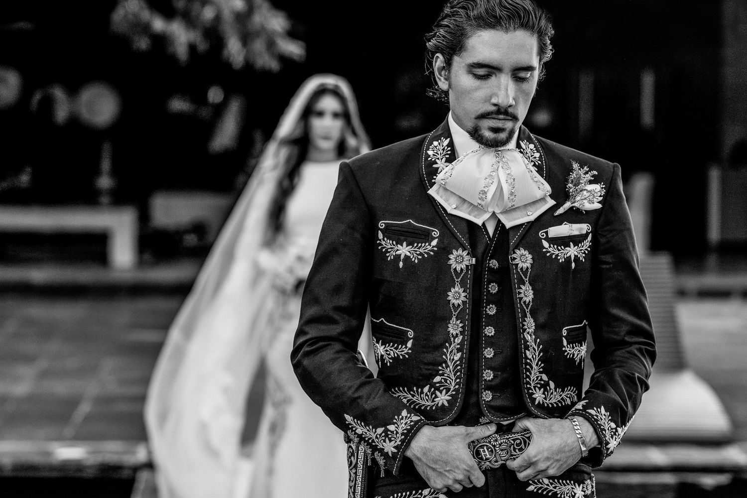 Black and white wedding photo of groom in ornate mariachi suit with bride in white gown blurred in background.