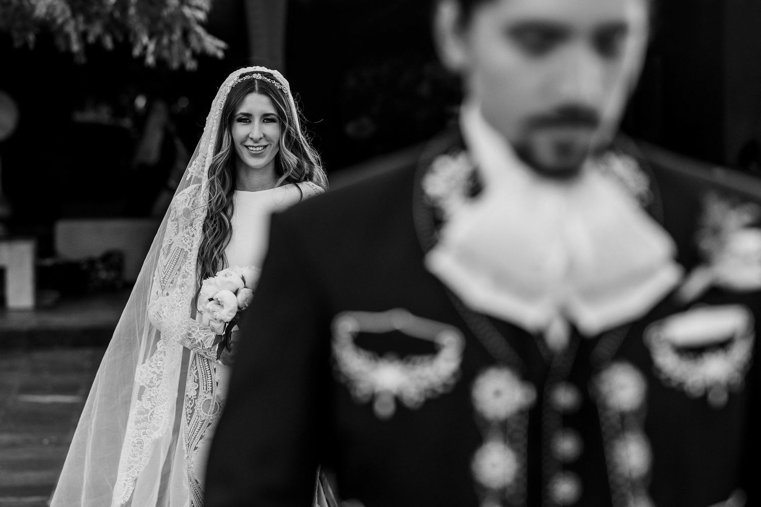Black and white wedding photo of smiling bride in lace veil walking toward groom in traditional charro suit.