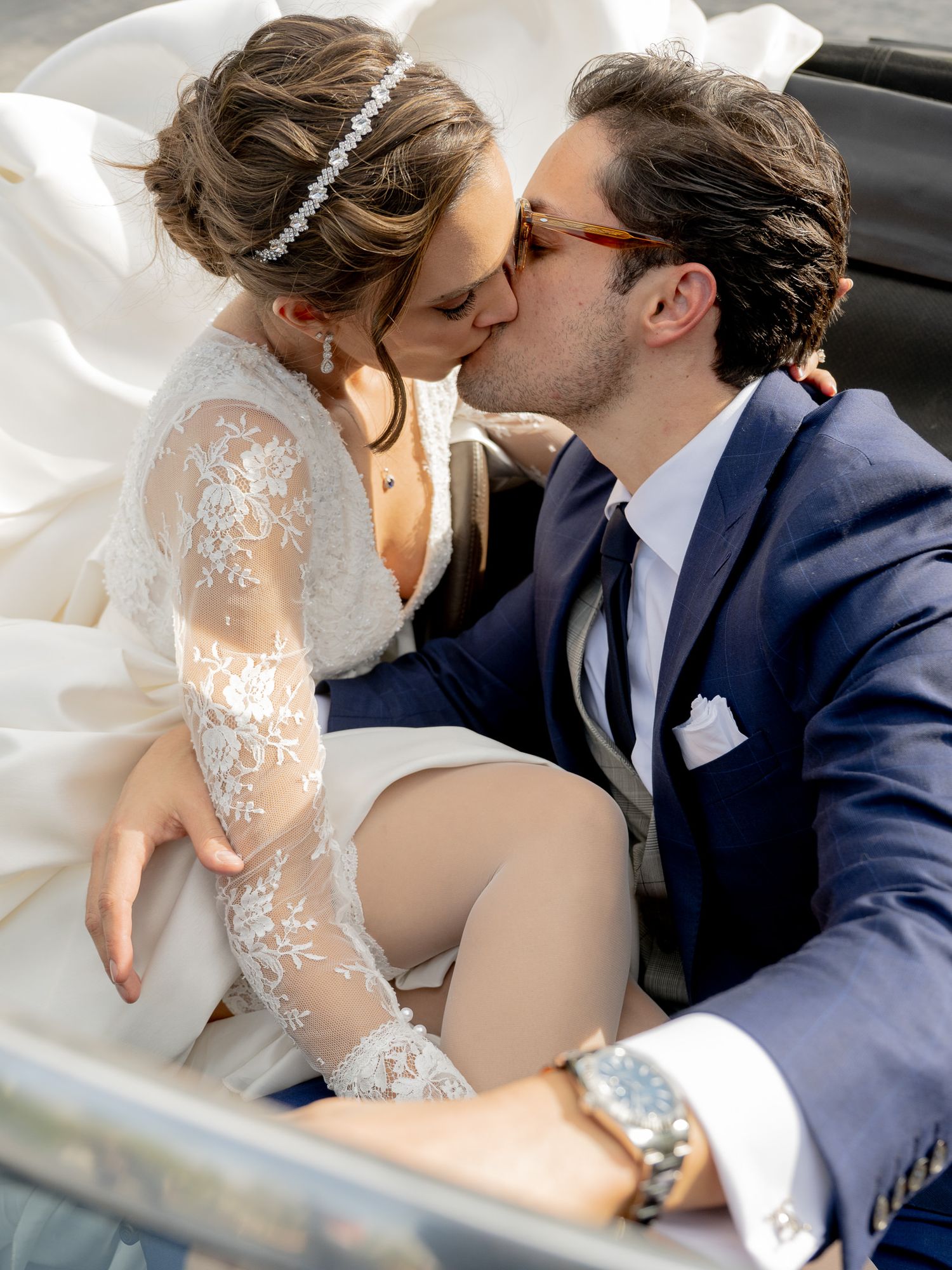 Bride and groom sharing a romantic kiss in a car, bride wearing lace wedding dress with headband.