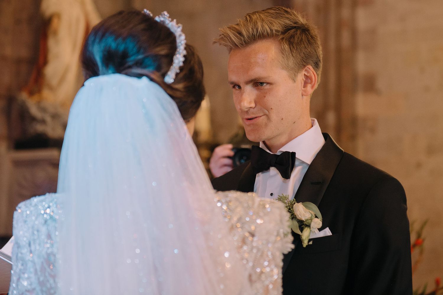 Groom in black tuxedo gazing at bride in sparkling white gown and veil during wedding ceremony.