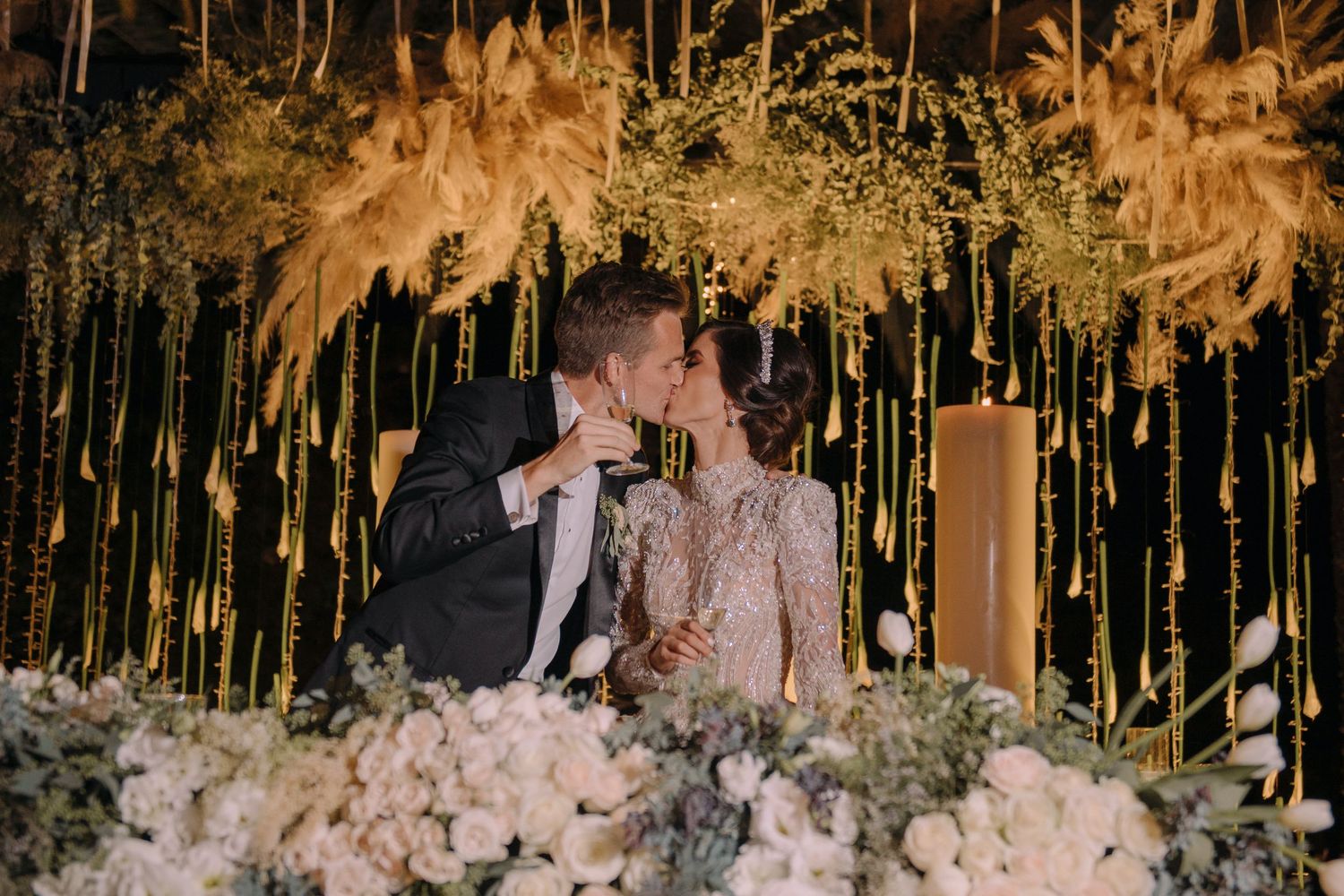 Couple sharing a romantic kiss at wedding reception altar adorned with lush floral arrangements and elegant hanging greenery.