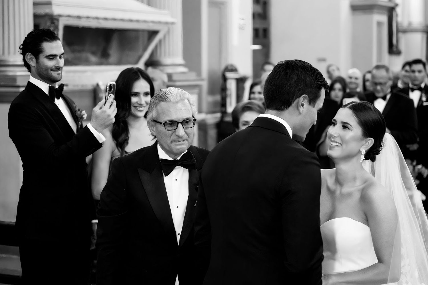 Black and white wedding ceremony photo with bride, groom, and guests smiling at elegant venue.