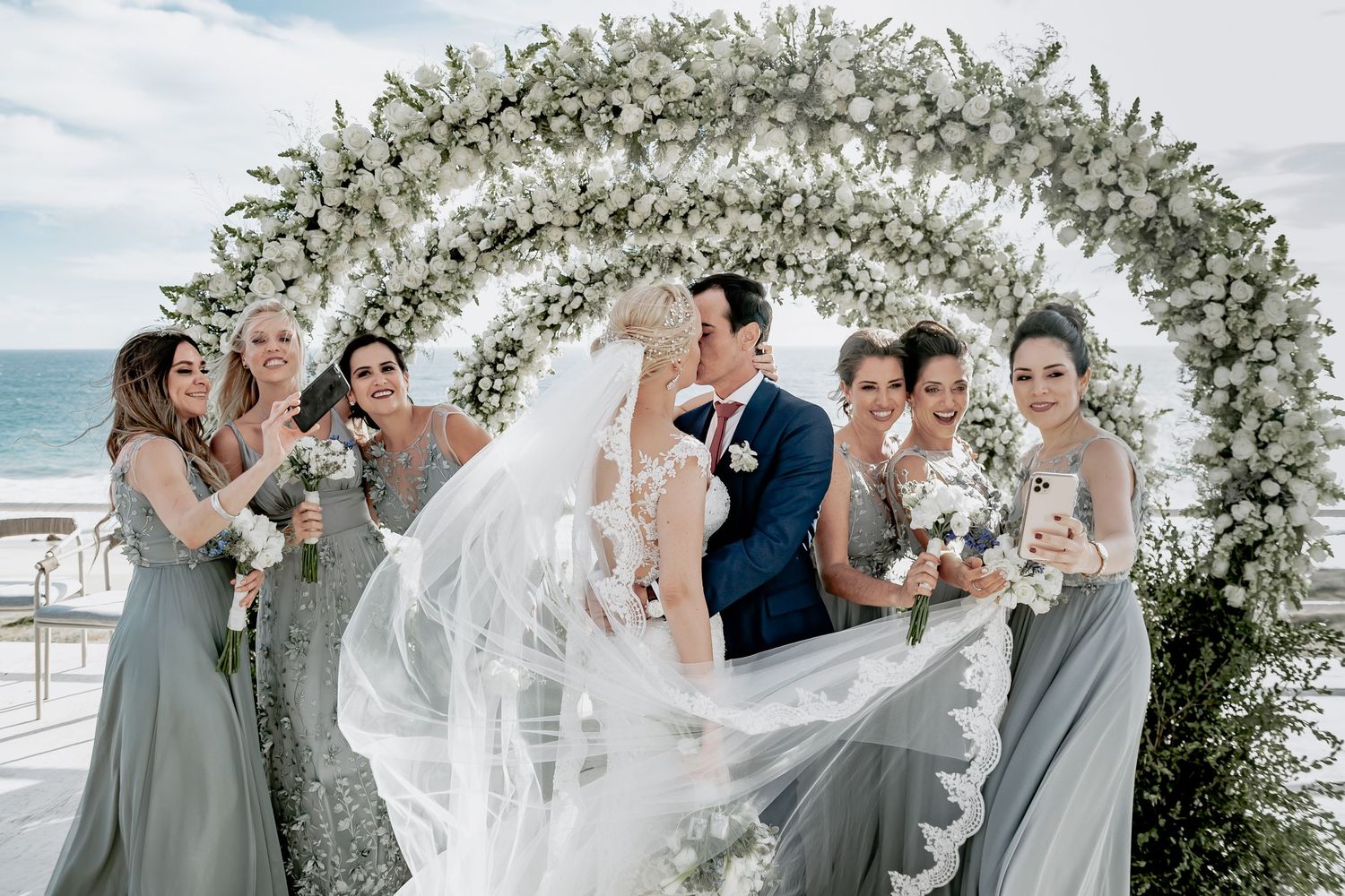 Wedding party celebrates under an elegant white floral arch by the ocean with bridesmaids in sage green dresses.