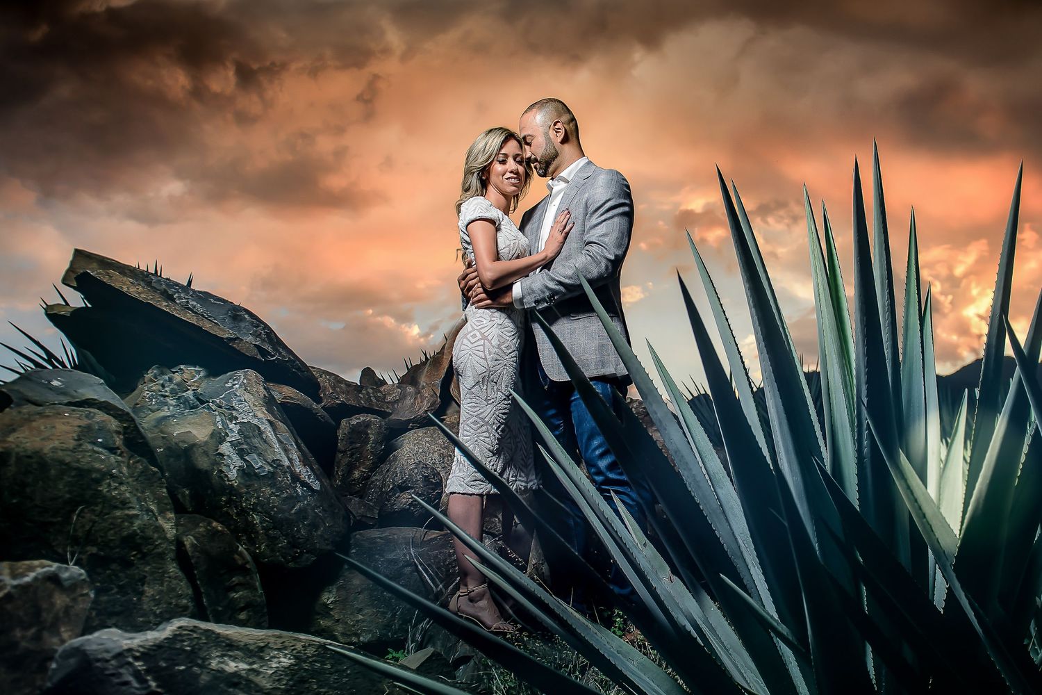 Couple embracing on rocks at sunset surrounded by agave plants under stormy sky.