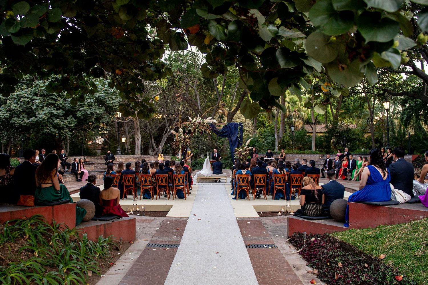 Outdoor wedding ceremony in a lush garden with guests seated in rows facing the altar on a white aisle runner.