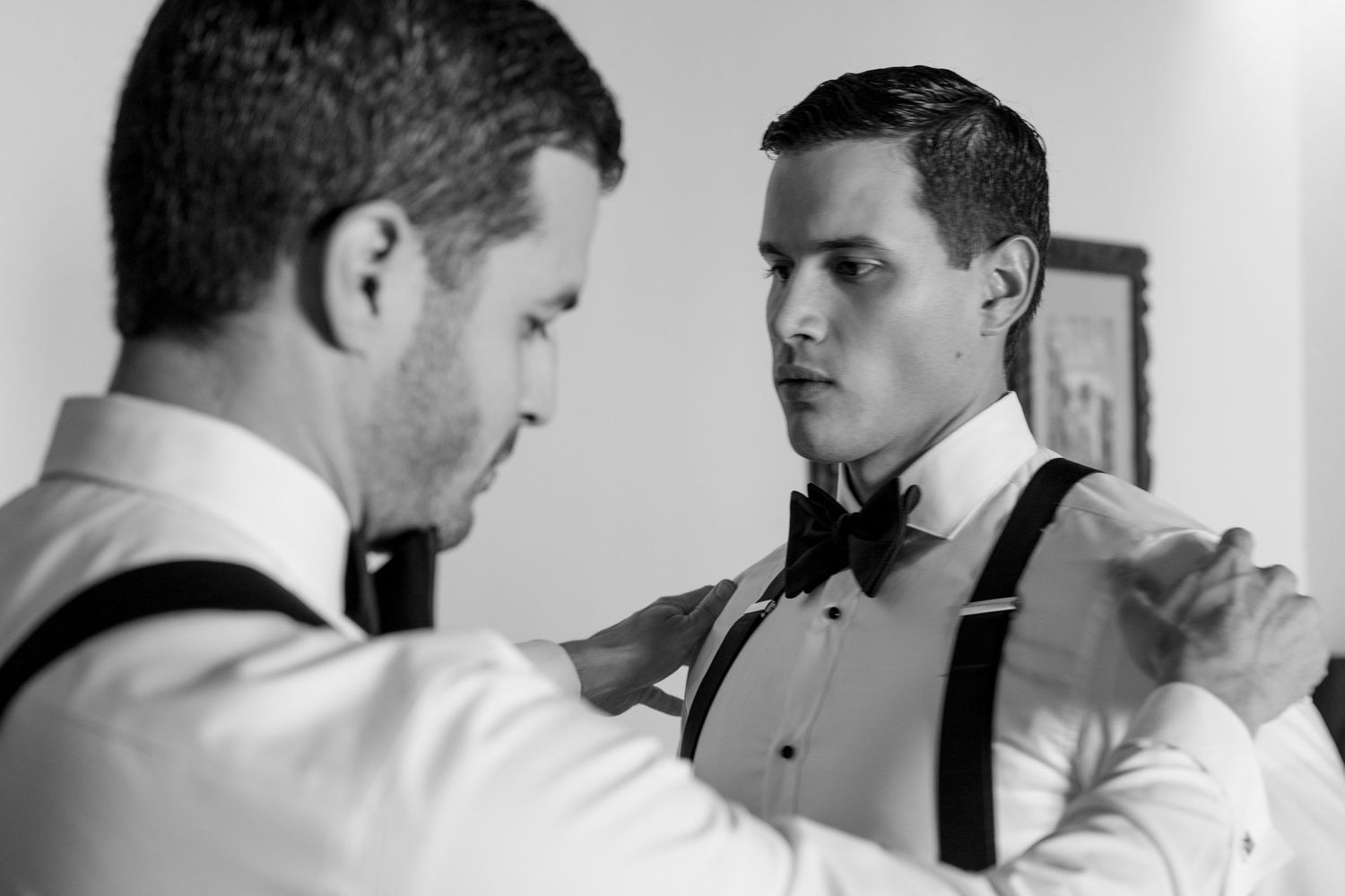 Two groomsmen in white shirts and suspenders adjusting bow ties in black and white wedding preparation photo.