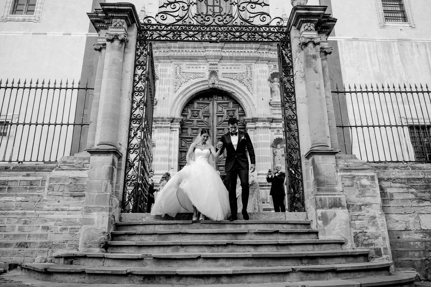 Black and white photo of a bride and groom descending stone steps outside an ornate church after their wedding ceremony.