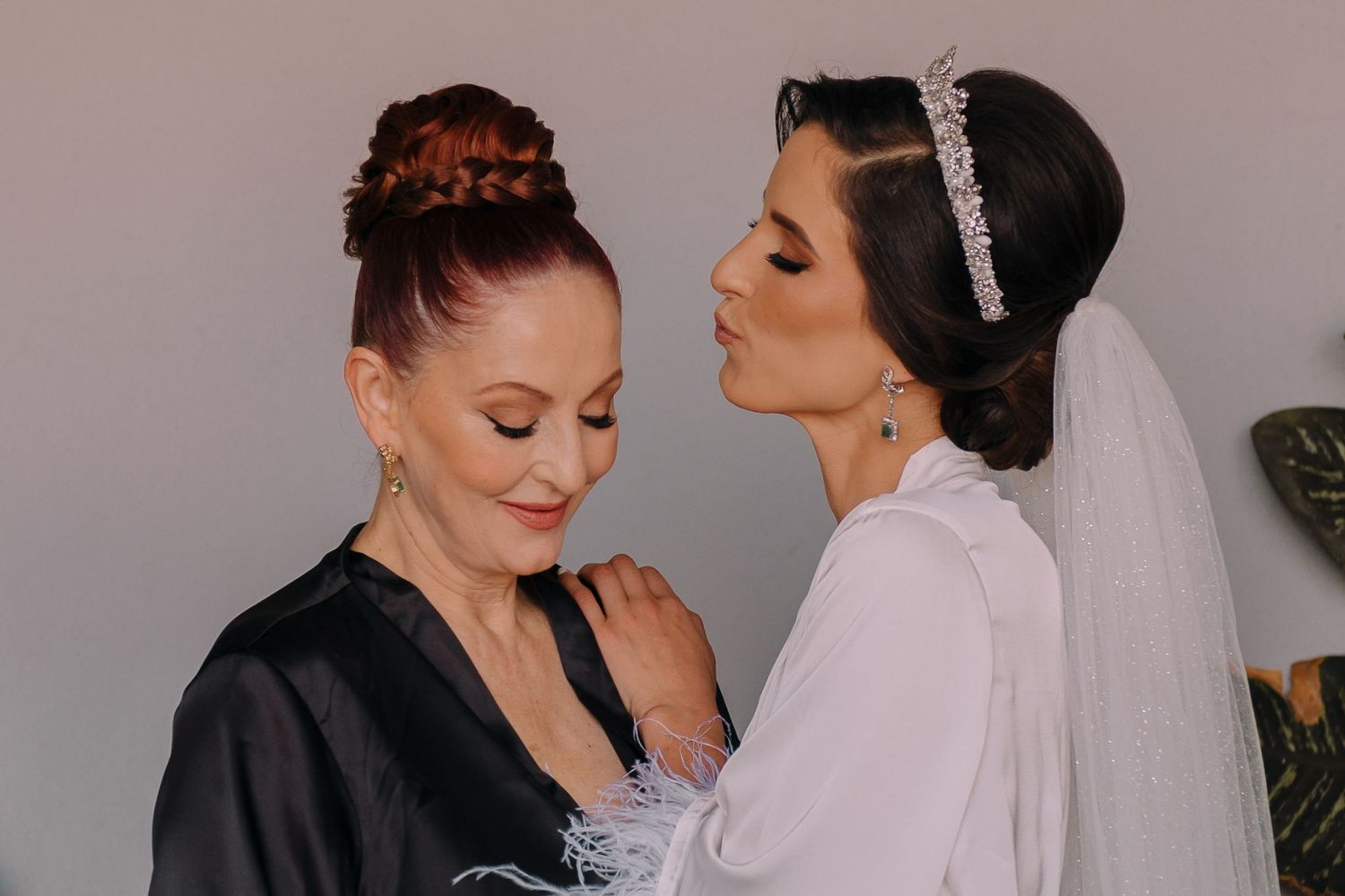 A bride in a white gown and veil kisses an older woman in a black robe during wedding preparations.