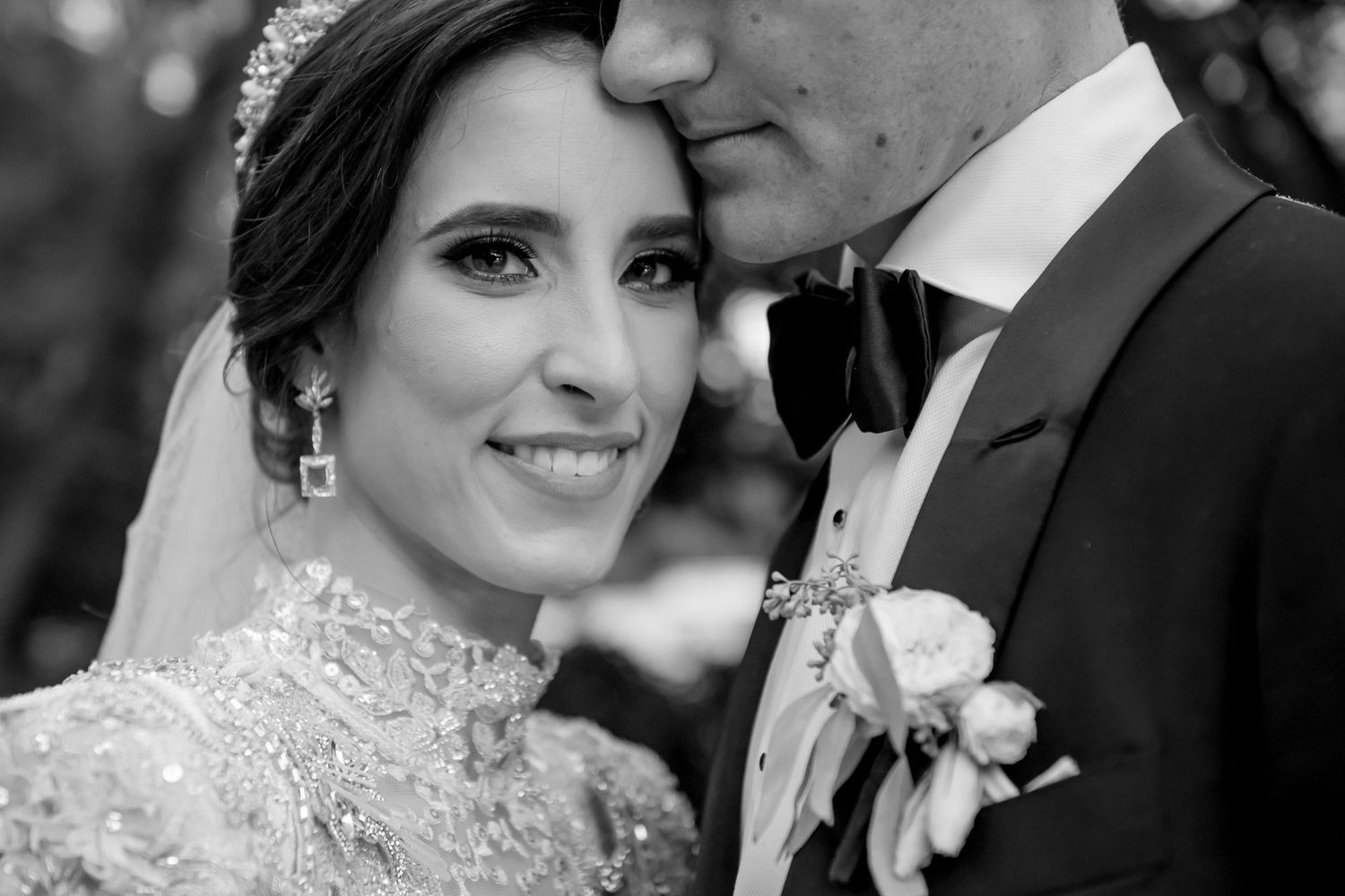 Black and white wedding portrait of smiling bride in beaded gown with groom in tuxedo kissing her forehead.