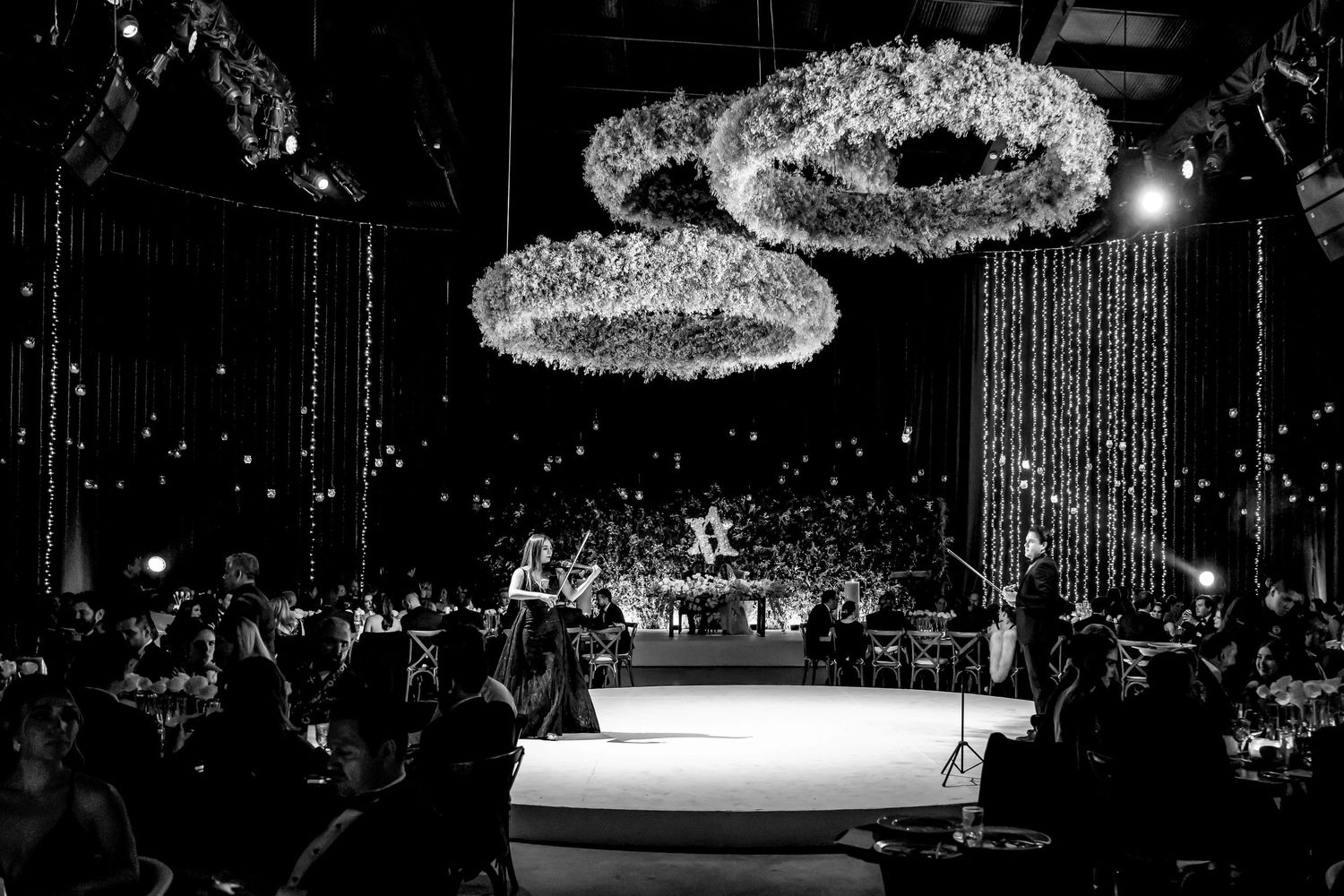 Elegant black and white wedding reception with floral chandelier rings, Star of David backdrop, and guests dining in grand hall.