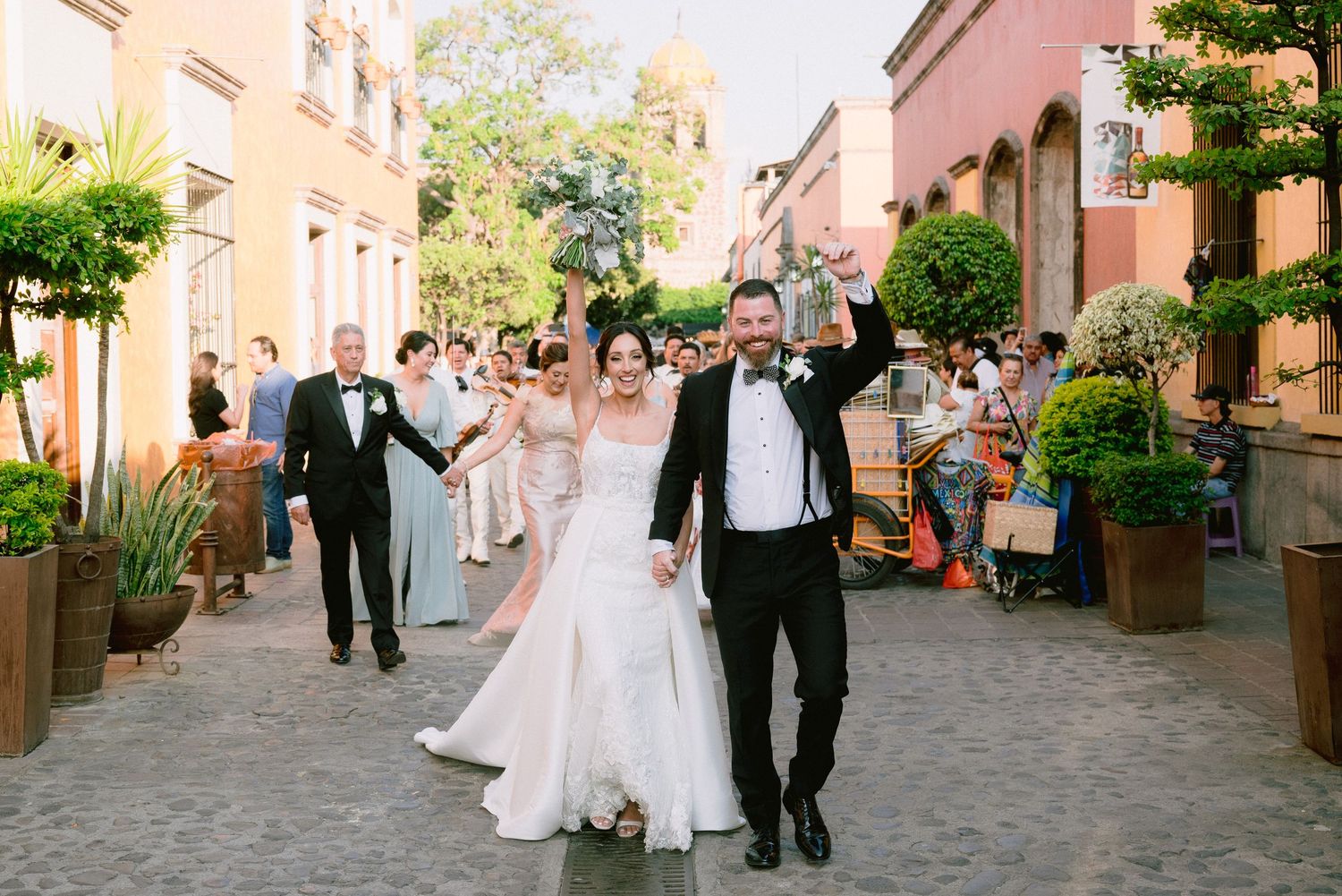 Bride and groom celebrate joyfully on a cobblestone street with wedding guests in formal attire in the background.