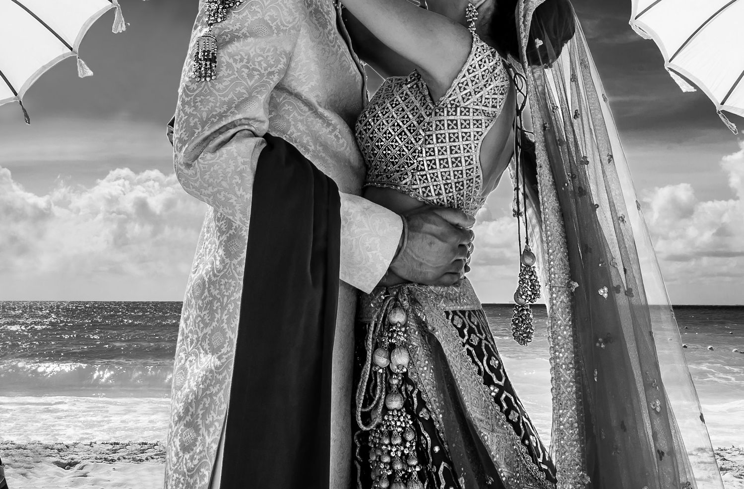 Black and white photo of a couple in traditional Indian attire embracing by the ocean under decorative umbrellas.