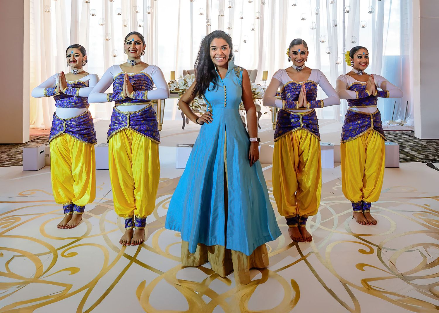 Woman in blue anarkali gown poses with four dancers in purple and yellow traditional Indian costumes.