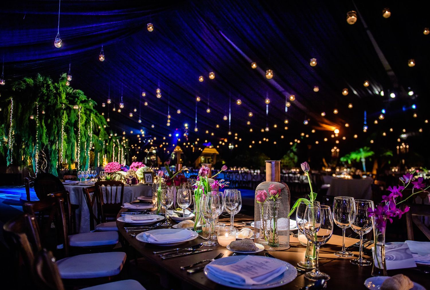 Elegant outdoor tent reception with string lights, lush greenery centerpieces, and colorful floral arrangements on dark wood tables.