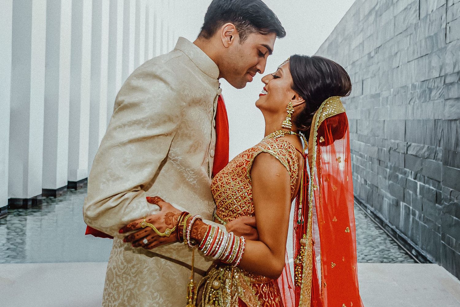 Couple in traditional Indian wedding attire embracing in a modern hallway with white walls.