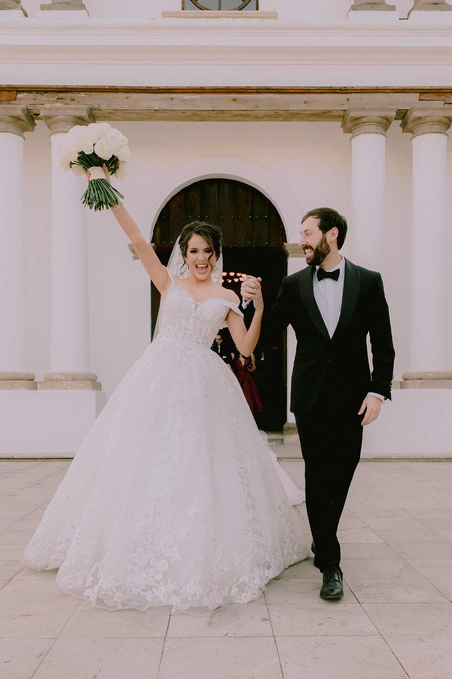 Joyful wedding couple exits venue with bride holding bouquet triumphantly in the air.